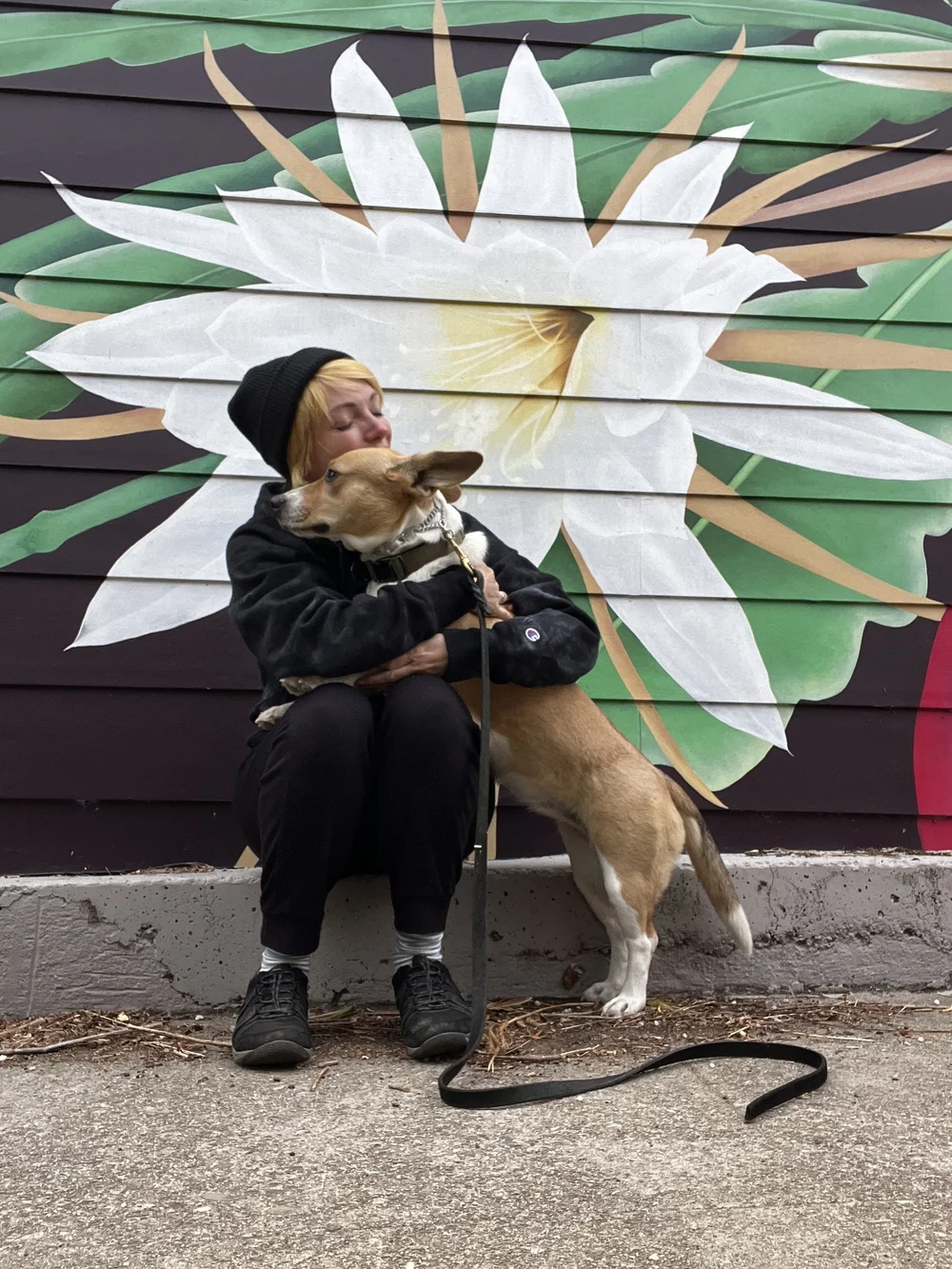 Kim of Good Company Canine sitting with her dog in front of a flower mural in San Francisco