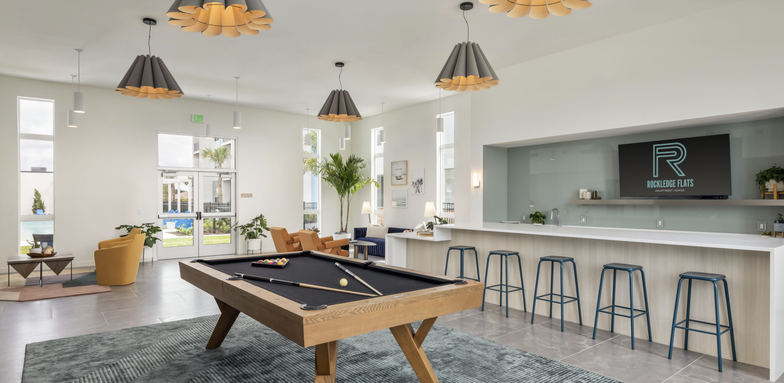 Interior of a modern lounge area with a pool table, bar seating, couches, and a TV displaying the logo of Rockledge Flats. The space has large windows, contemporary lighting, and decorative plants.