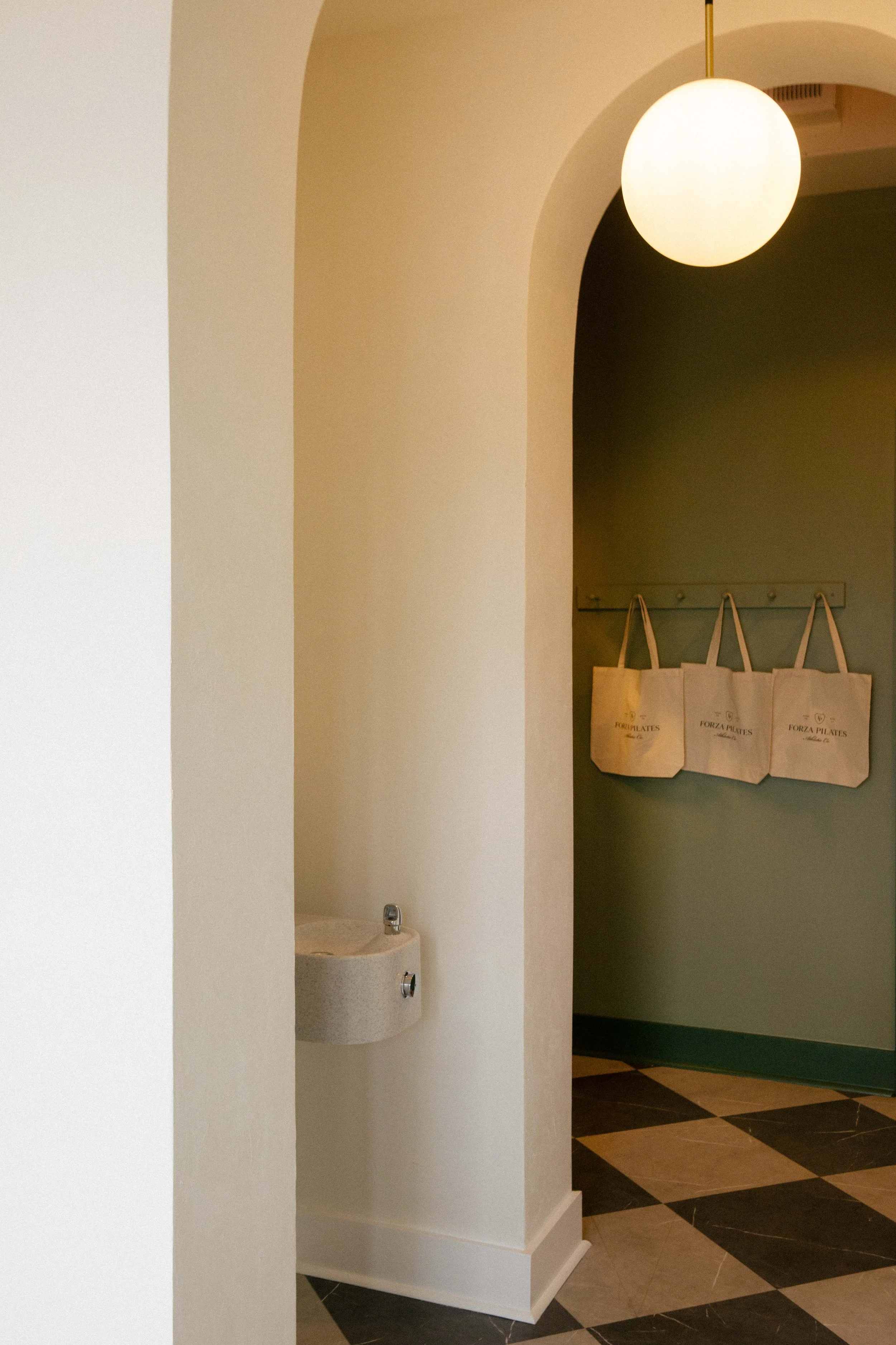 Interior of a room with beige walls, a white hanging spherical light, a small wall-mounted sink, and three tote bags hanging on hooks.
