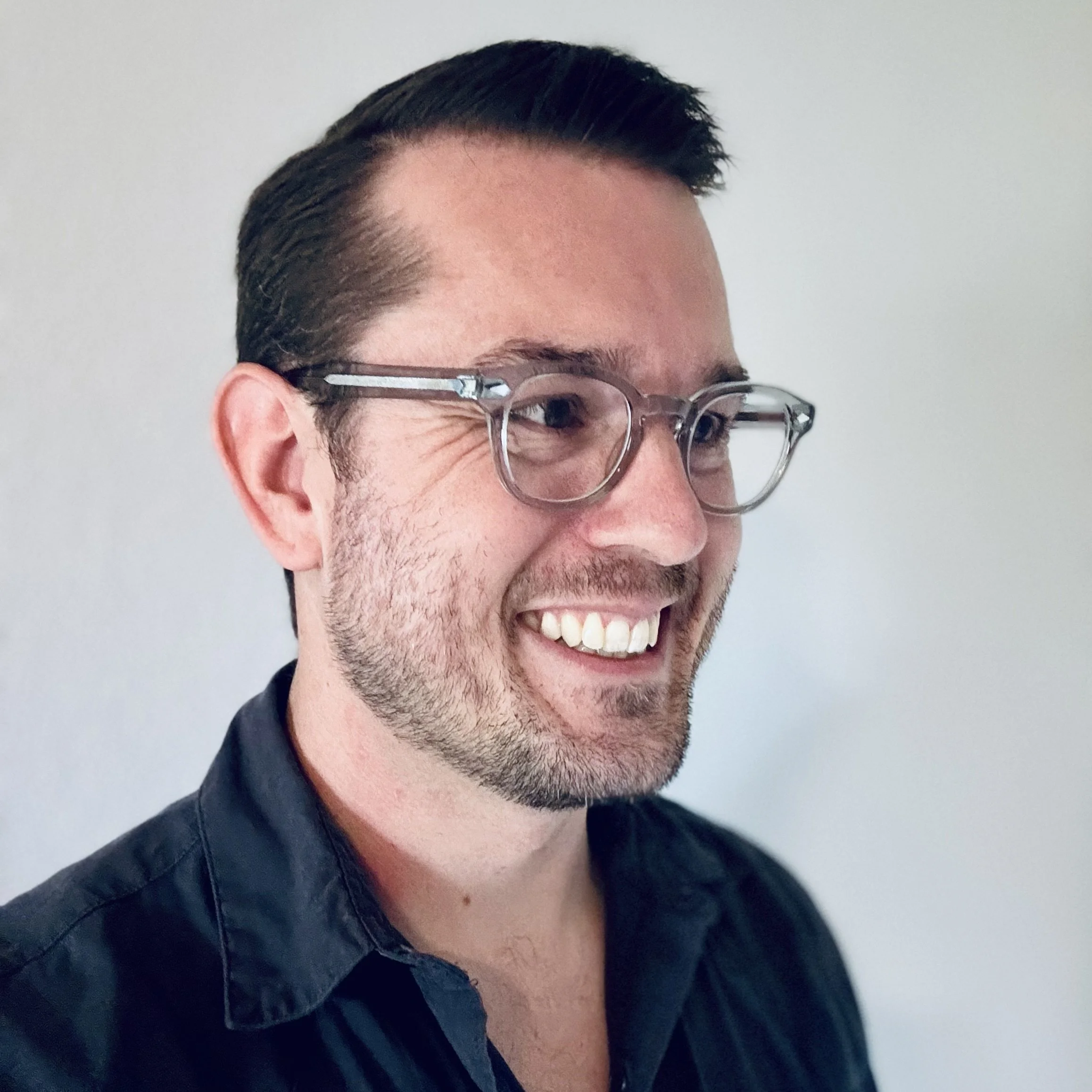 Close-up of a smiling man with glasses, short dark hair, and wearing a dark jacket, against a plain background.