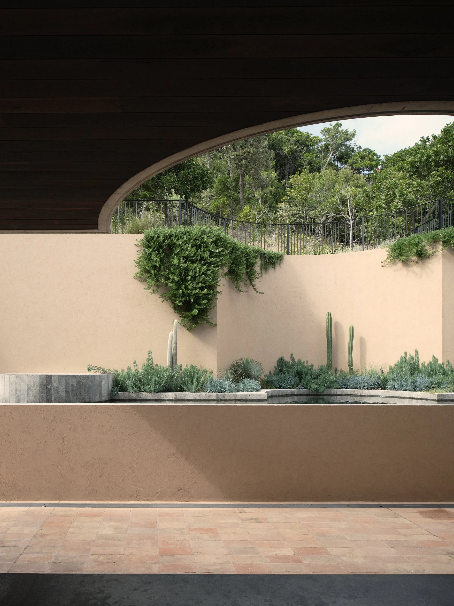 A modern architectural view of an outdoor space with a beige wall, desert plants, and trees in the background.