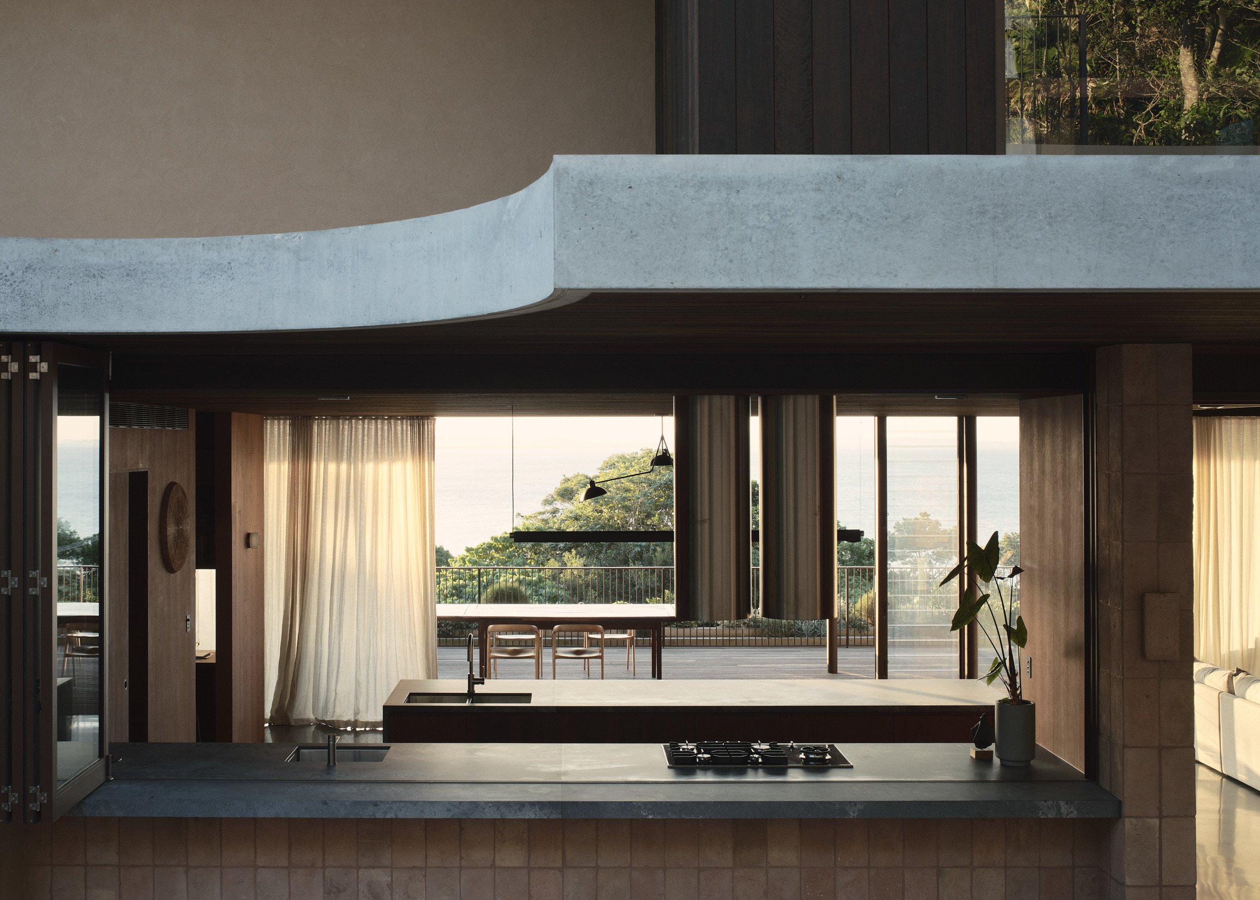 Modern kitchen with large open windows, view of trees and water, kitchen island with sink, and curtains.