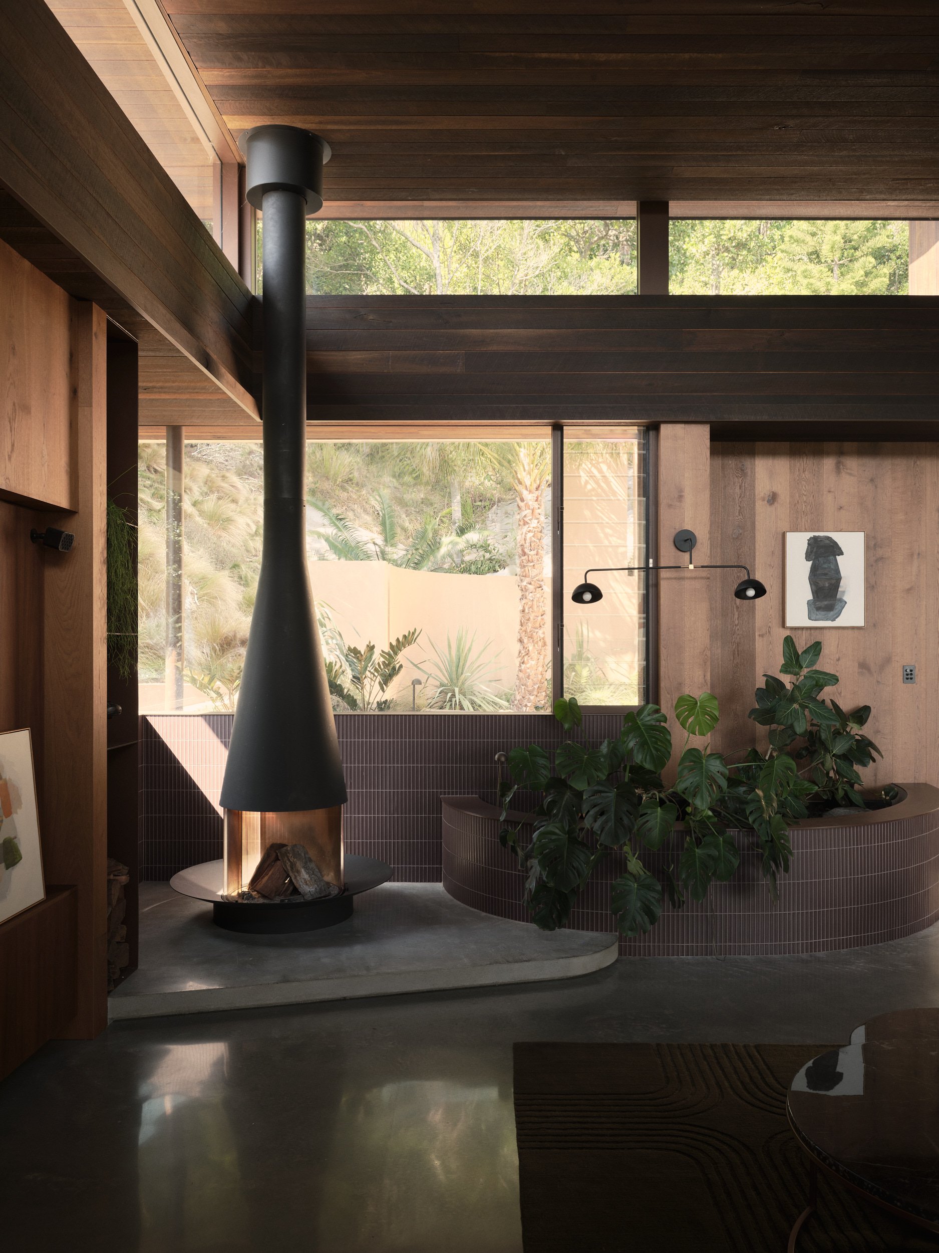 Interior of a modern house with a wood ceiling, large windows showing trees outside, a black conical fireplace with logs, indoor plants, and minimalistic décor.