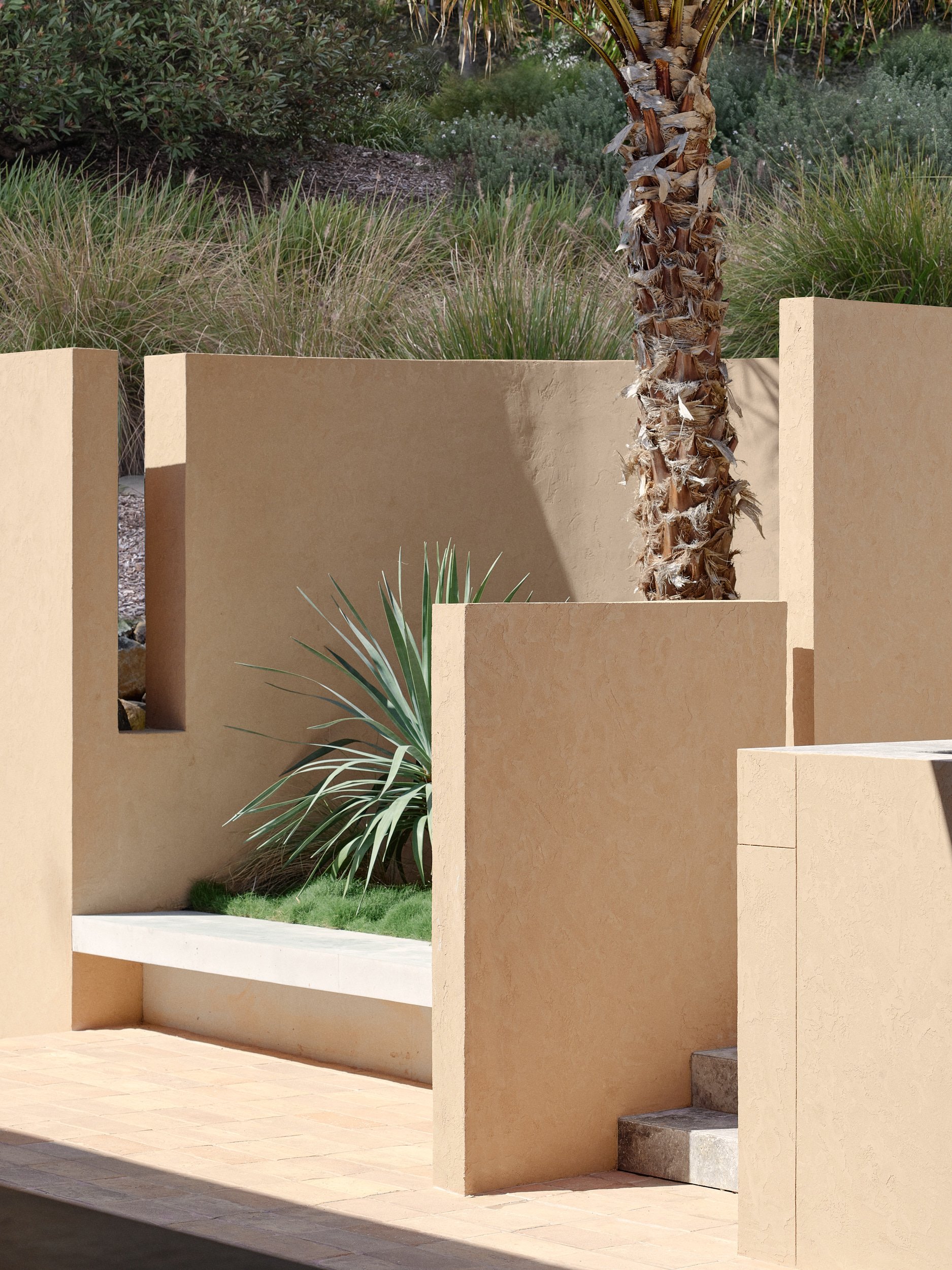 A modern outdoor space featuring beige stucco walls, a palm tree, and desert plants in the background, with a minimalist aesthetic.