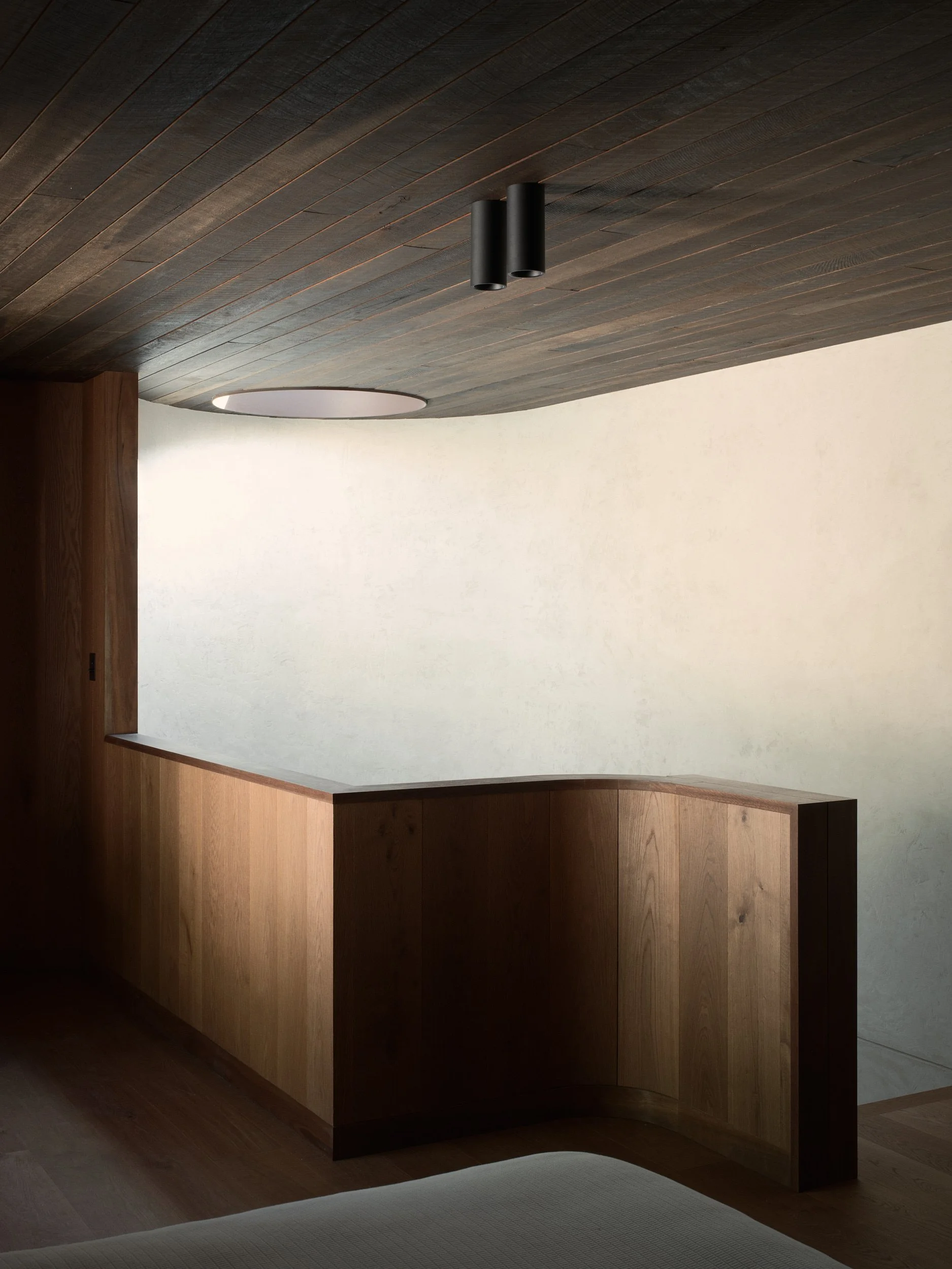 Interior of a room with a dark wooden ceiling, beige wall, wooden furniture, and a corner of a white bed or sofa.