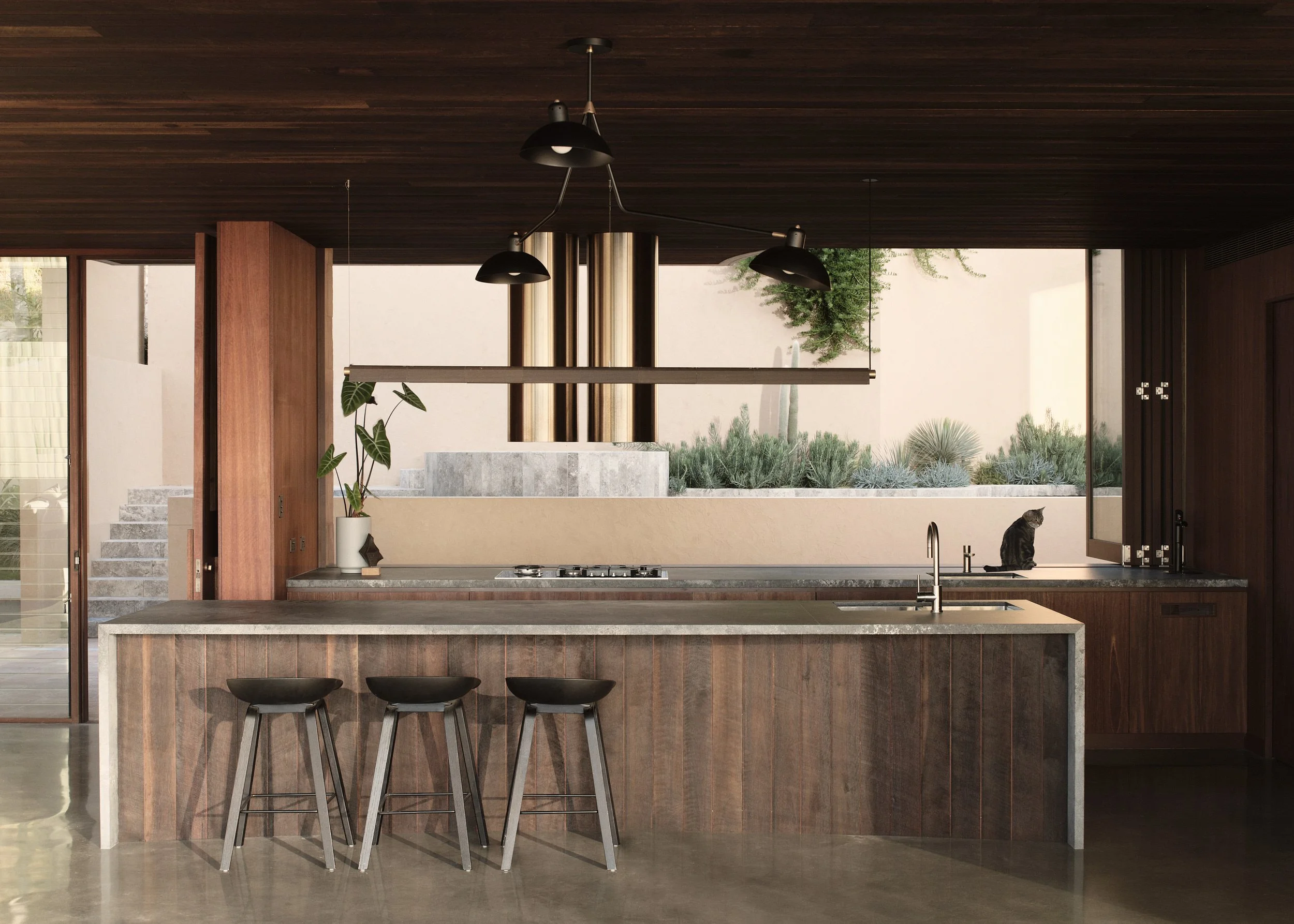 Modern kitchen with dark wood cabinetry and a large window overlooking a desert landscape with plants. The kitchen features a marble island with three black stools, a sink, and hanging black pendant lights.