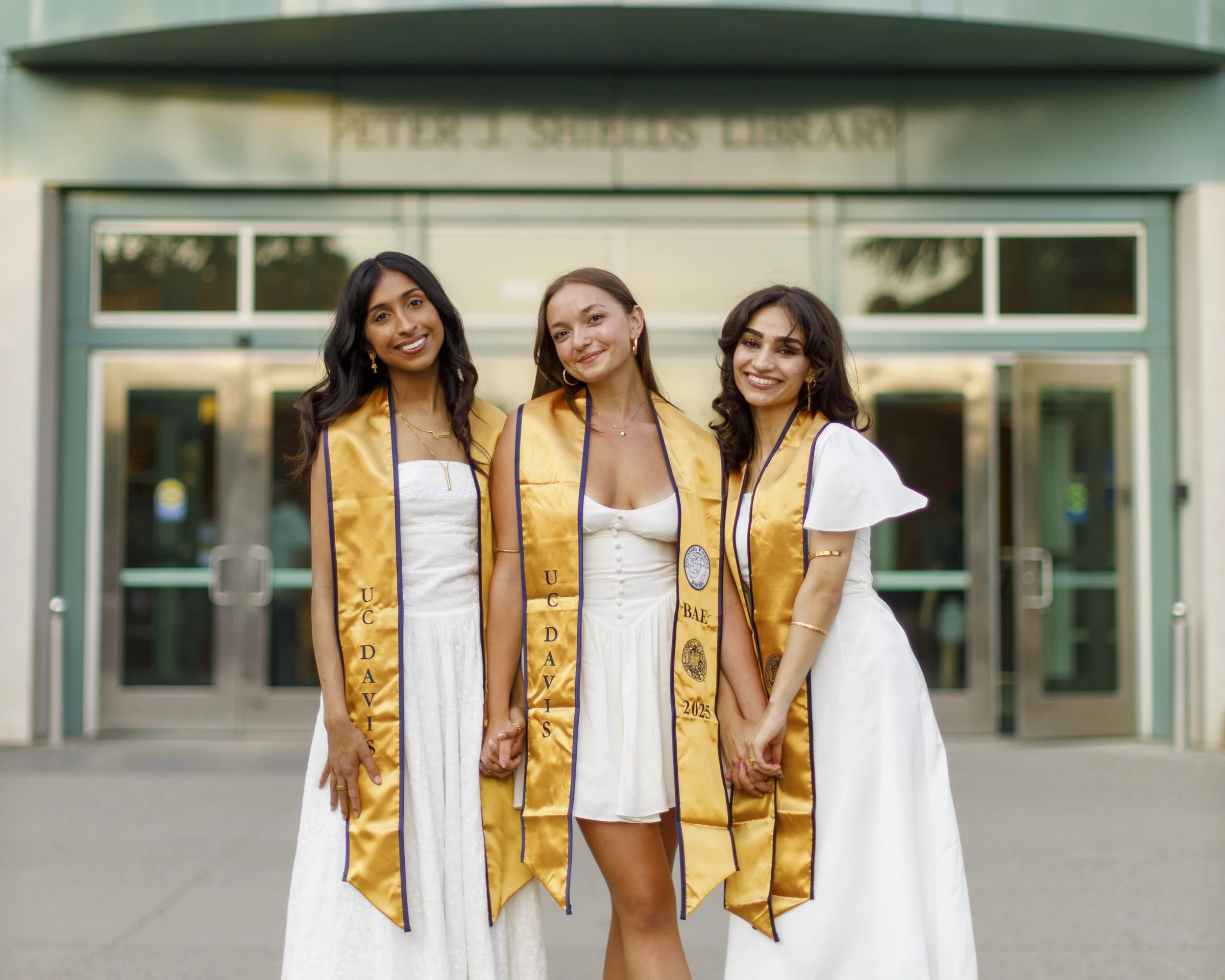 Three women in white dresses and gold graduation stoles smiling outside of Shields Library at UC Davis.