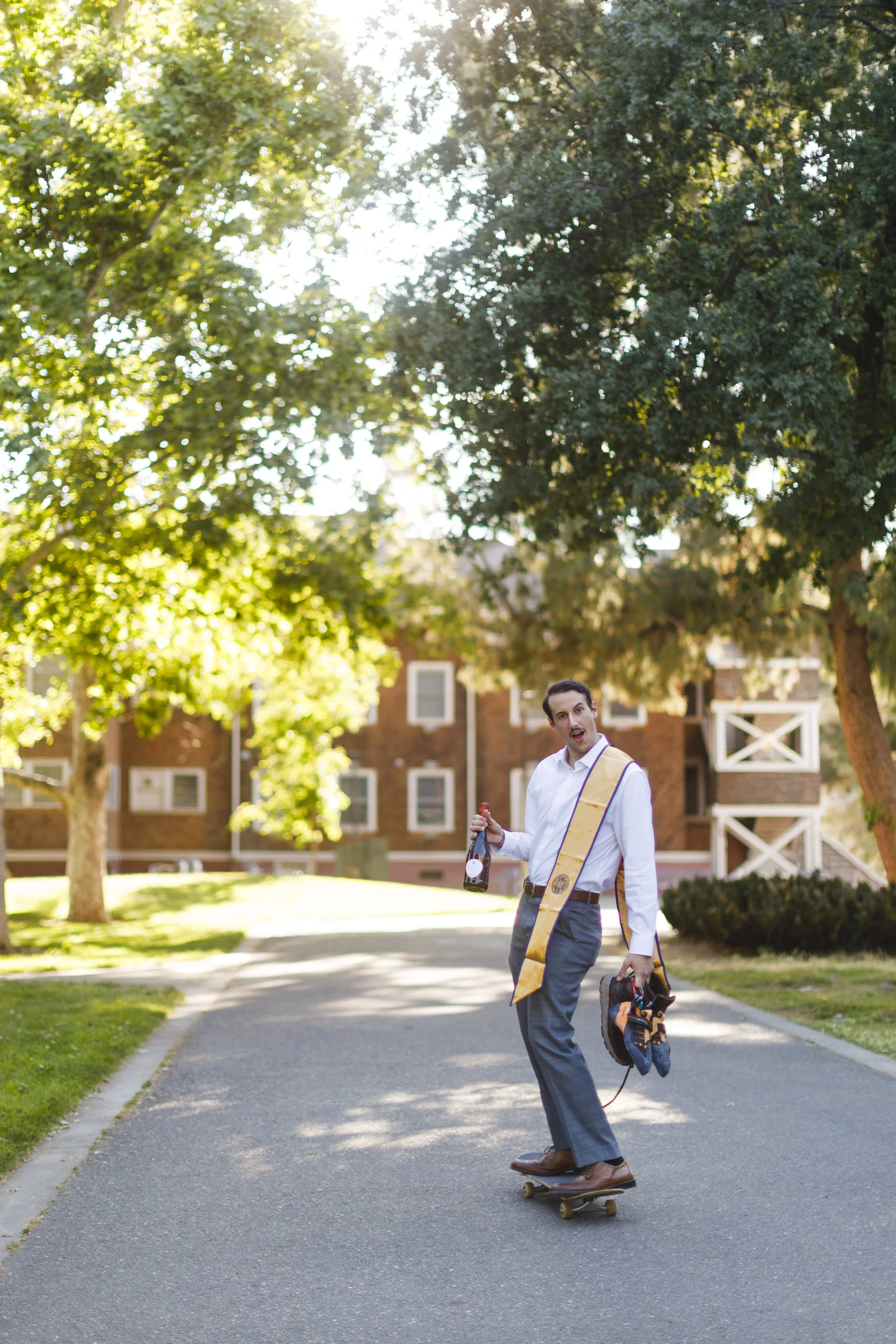 Fun solo grad photo of a young man on a skateboard holding champagne, rock climbing shoes, and hiking boots.