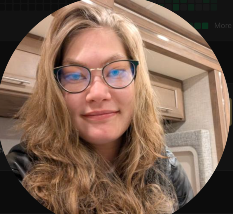 A woman with long wavy hair wearing glasses, smiling, in a setting with wooden cabinets and a light-colored wall.