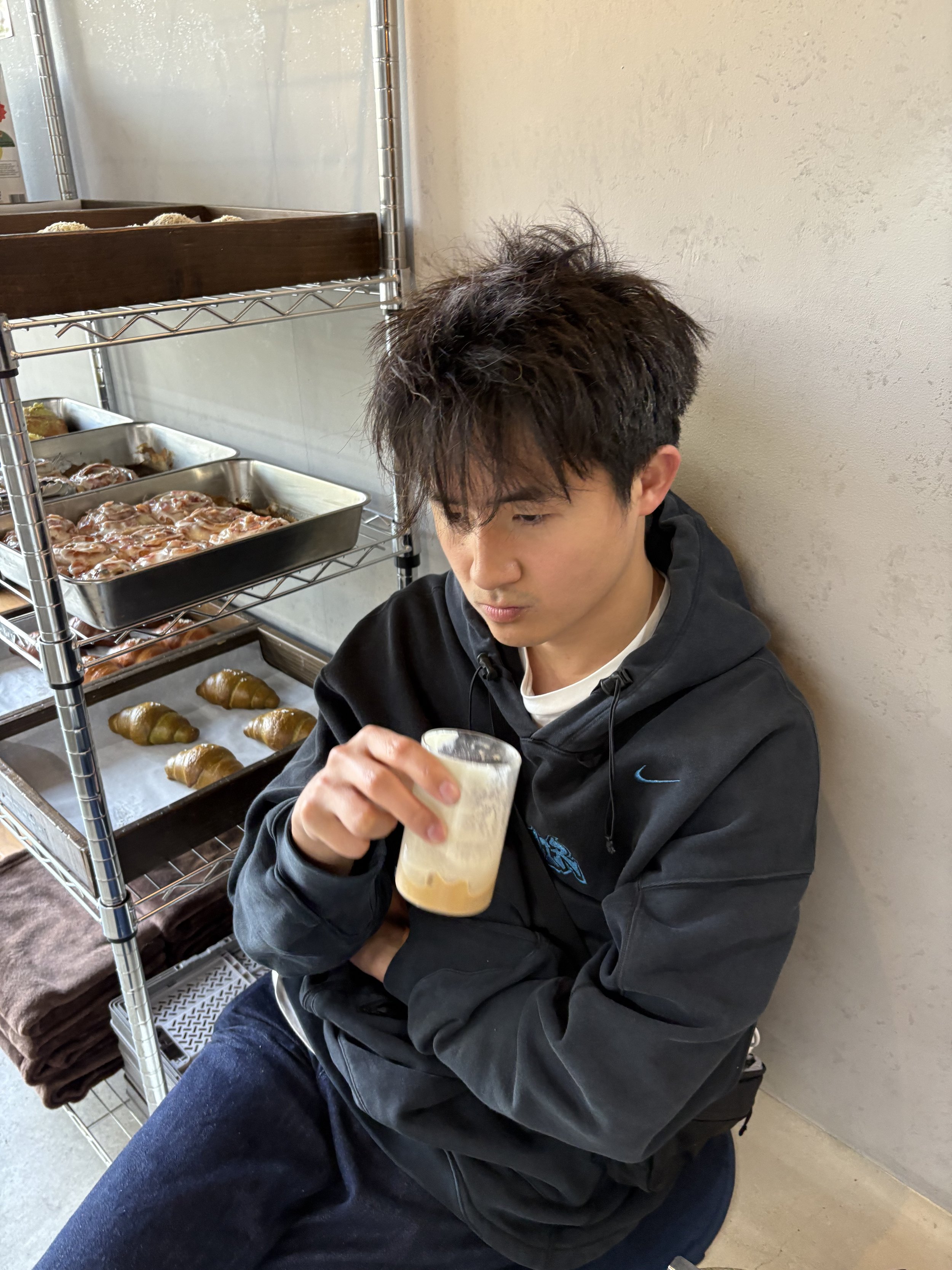 Young man with messy hair wearing a black hoodie sitting against a wall, holding a plastic cup with a light-colored drink, next to a metal shelving unit with baked goods like croissants and pastries.