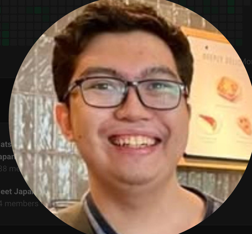A young man with glasses smiling in an indoor setting, with a blurred background featuring a display board.