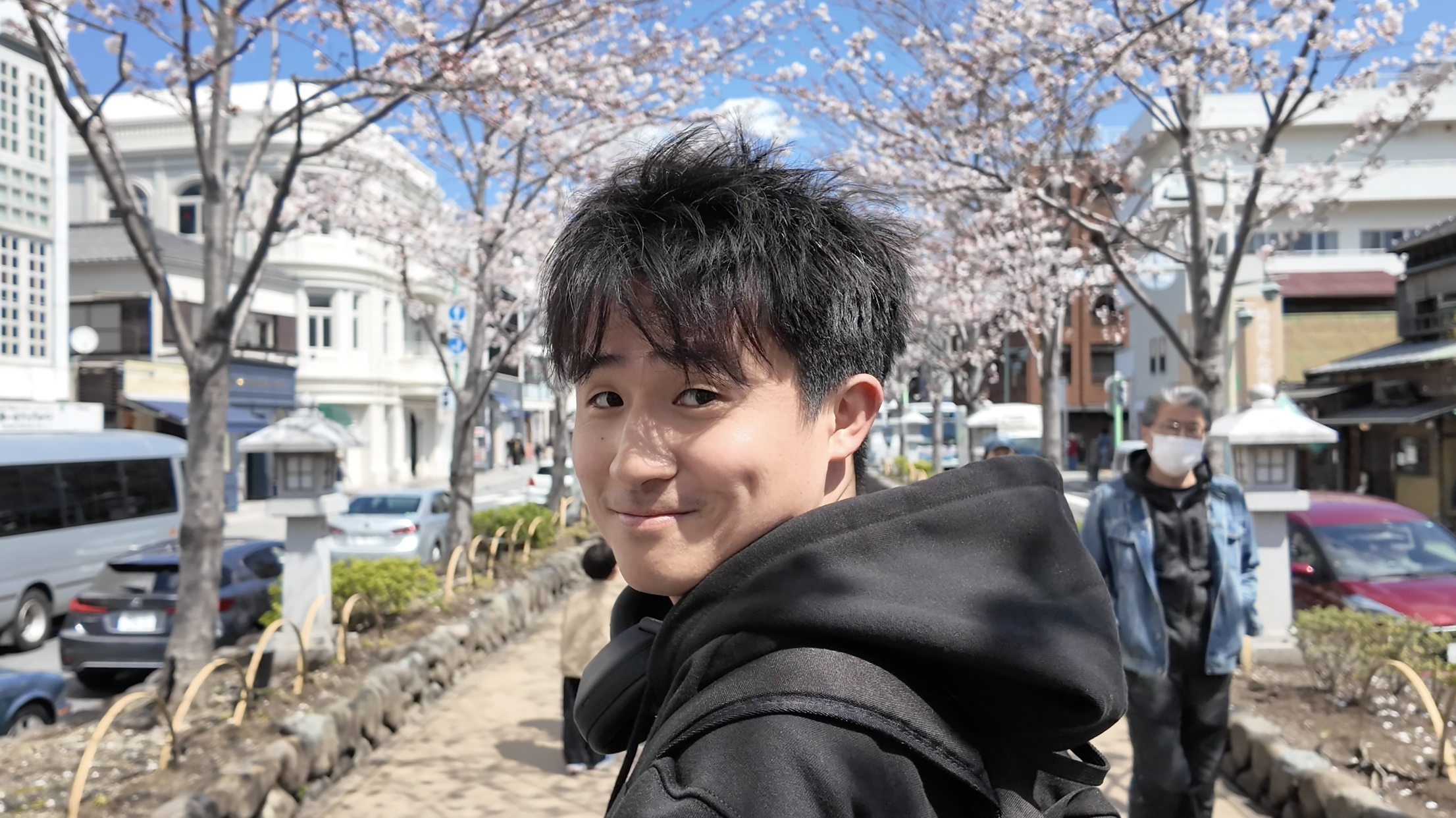 Young man with dark hair smiling on a city street lined with blooming cherry blossom trees, with people and cars in the background.