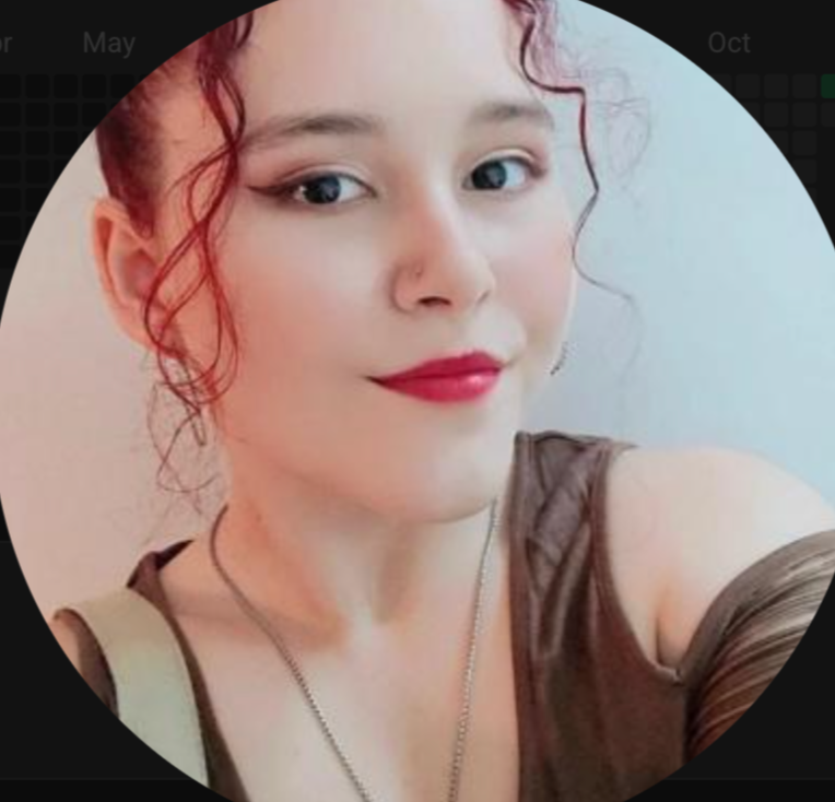 A woman with red, curly hair and fair skin, wearing red lipstick, a brown sleeveless top, and a necklace, posing for a selfie against a neutral background.