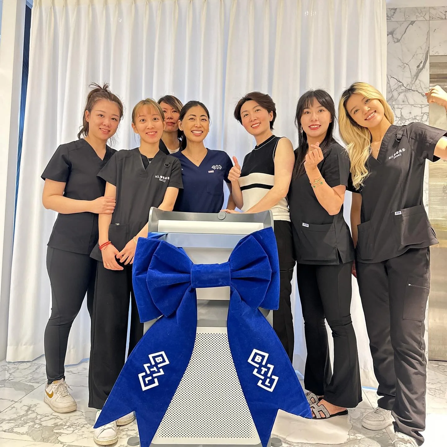 Group of seven women posing together around a decorated object with a large blue bow with white patterns in a room with white curtains and marble flooring.