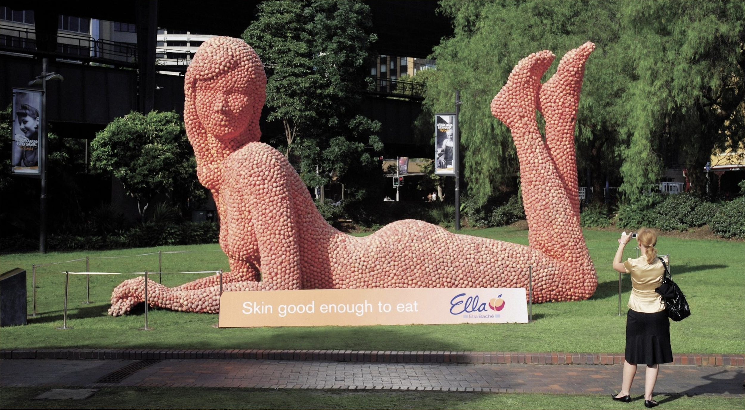Large sculpture of a reclining woman made from peaches, with a sign reading "Skin good enough to eat" by Ella Baché, in a park setting. A person is taking a photo of the sculpture.