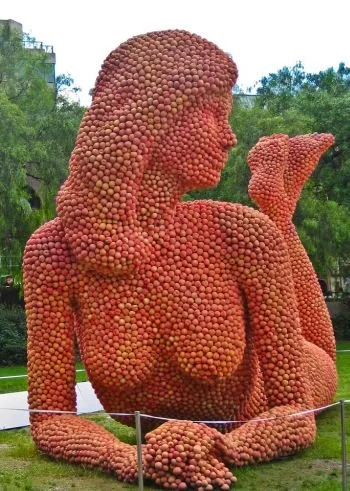 Sculpture of a reclining female figure made from apples on display in a park.