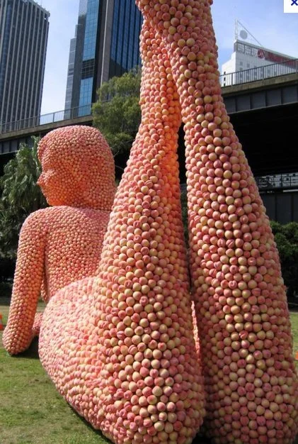 Sculpture of a reclining figure made of apples with city buildings in the background.