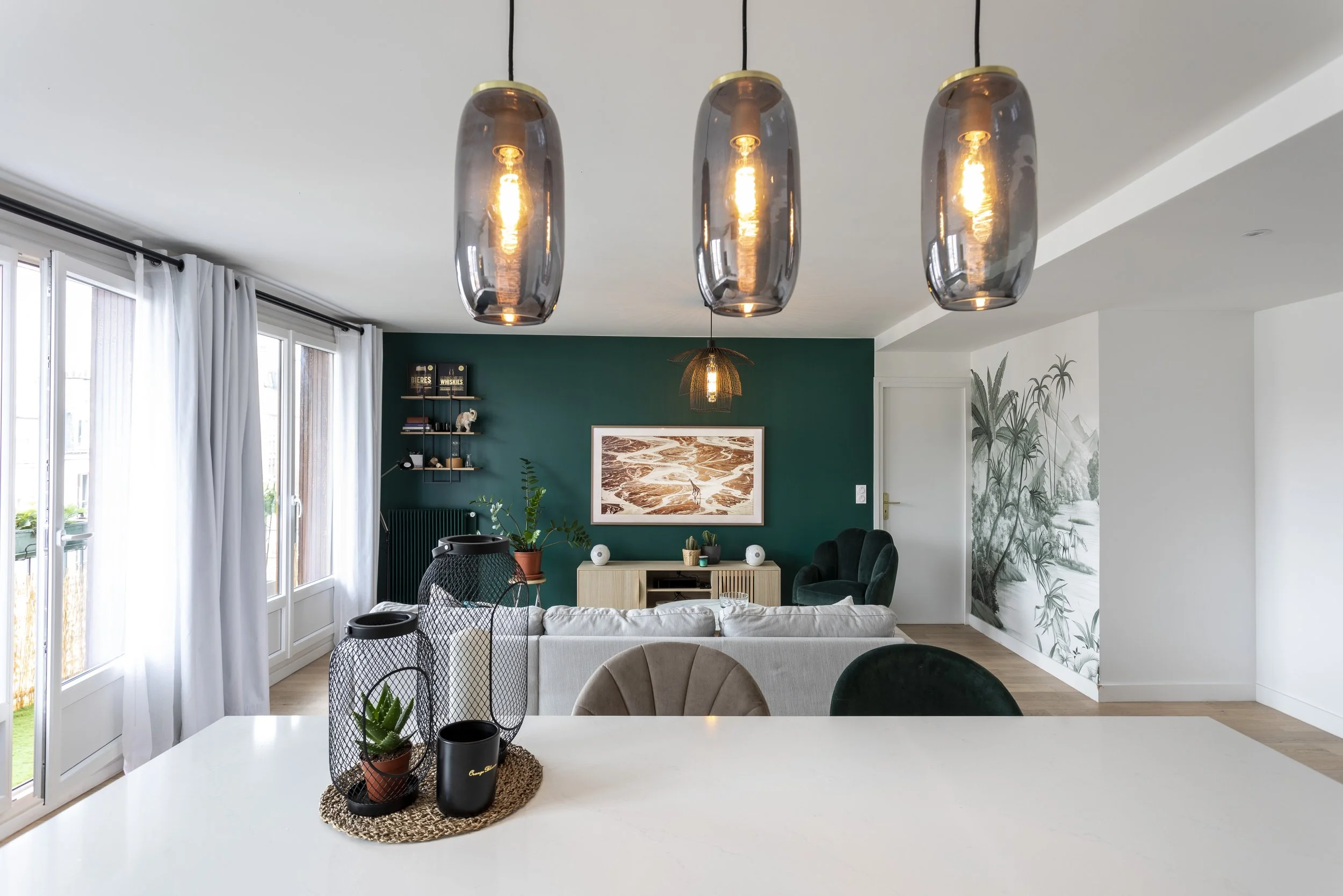 Salon moderne avec mur vert foncé, canapé beige, fauteuil vert, peinture abstracte, étagère noire avec décorations, et grand mur avec photo de nature. Lampes suspendues en verre foncé au plafond.
