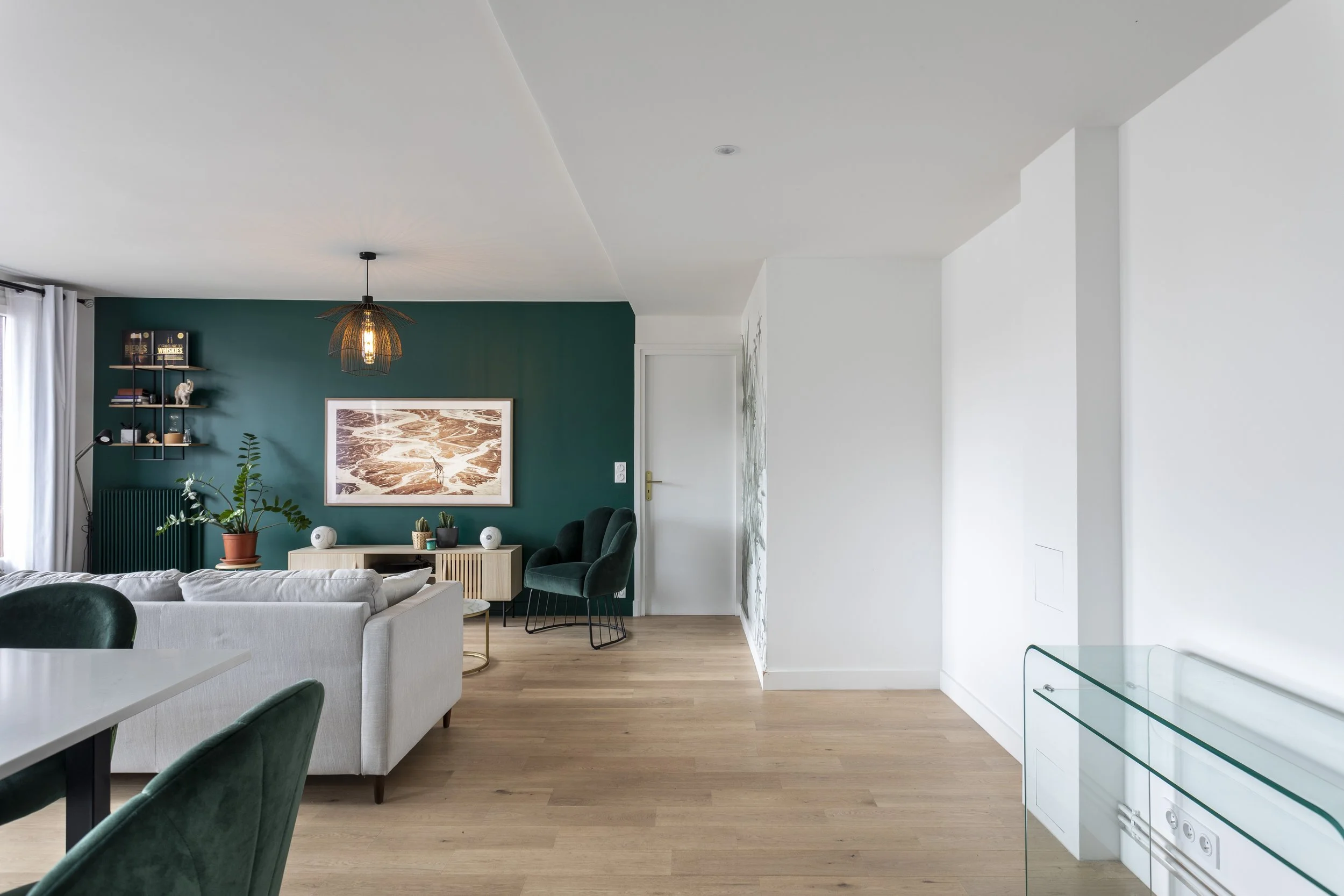 Salon moderne avec mur vert foncé, canapé beige, plantes vertes, tableaux, étagères et meubles en bois, parquet en bois clair.