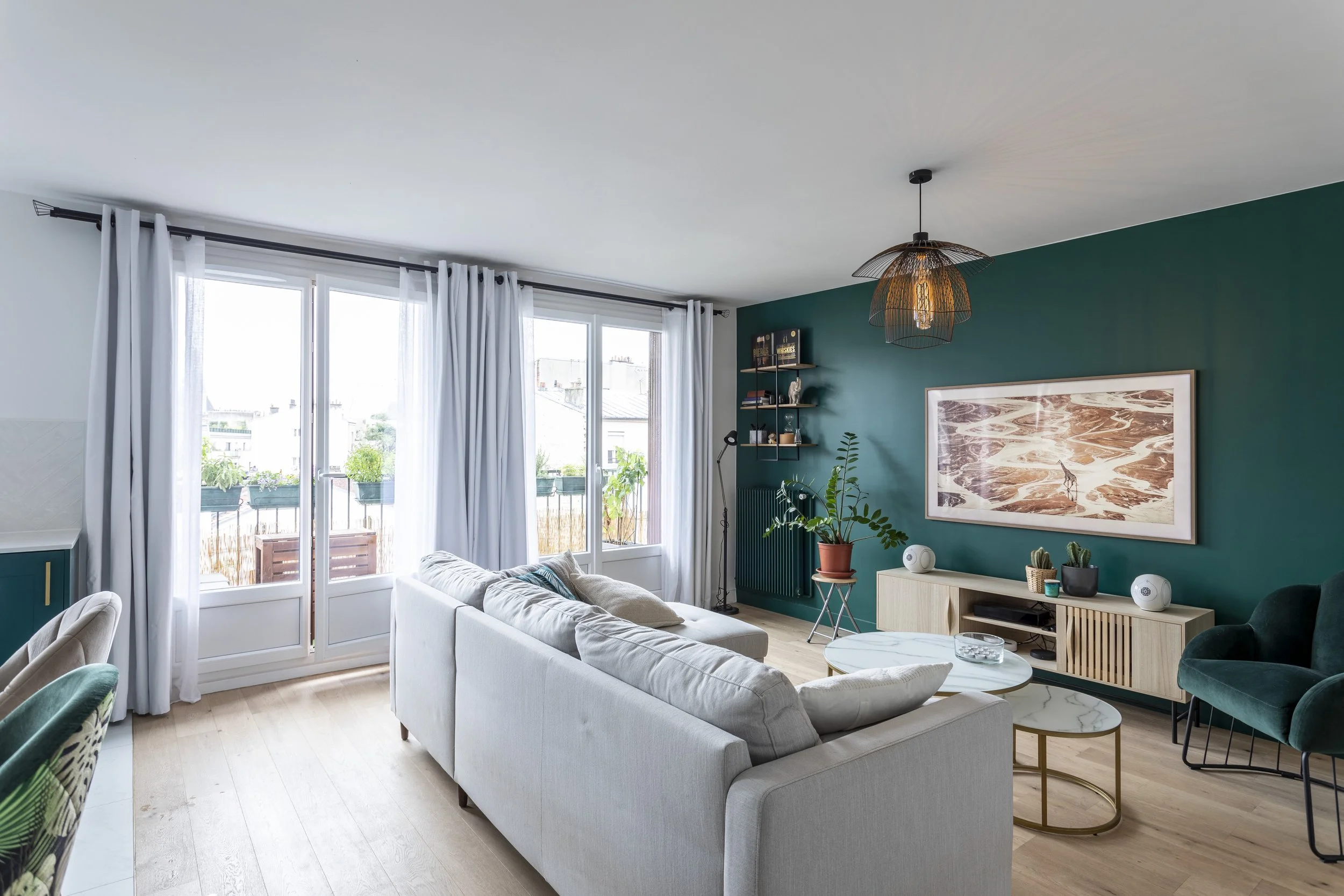 Salon contemporain avec mur vert foncé, canapé beige, table basse ronde, fauteuil vert, étagère murale avec livres et décorations, grande fenêtre avec rideaux blancs, plantes d'intérieur