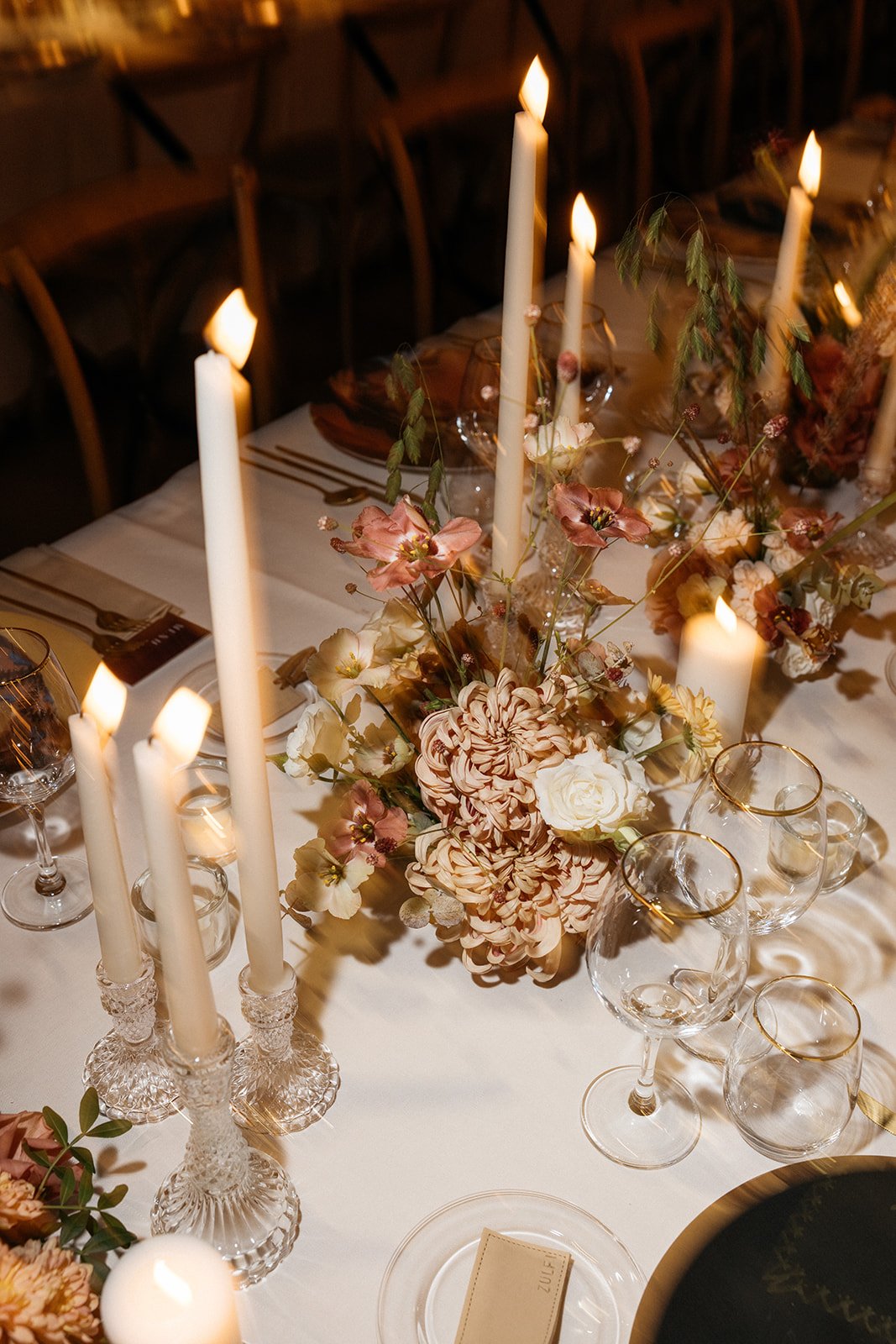Elegant dining table with tall white candles in crystal holders, a floral centerpiece with large pink, white, and cream flowers, and gold-rimmed wine glasses.
