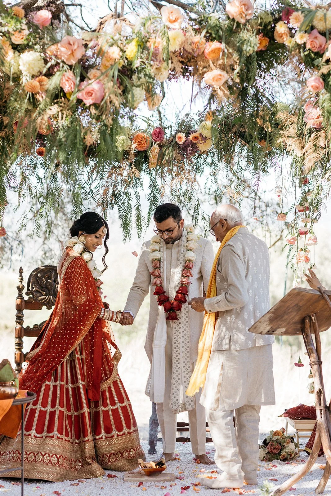 A couple in traditional Indian red wedding attire participating in a wedding ceremony, holding hands, under a floral canopy, with an officiant and an elderly man.