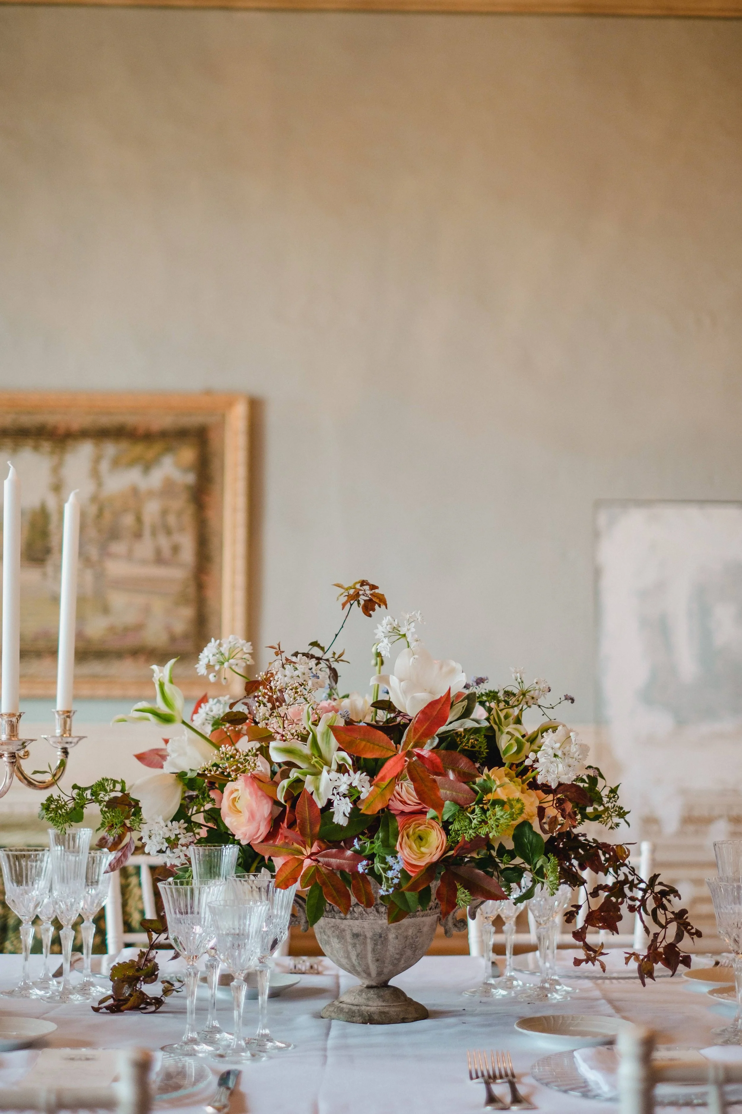 Elegant dining table with a large floral centerpiece in a decorative vase, surrounded by glassware, plates, and silverware, in a decorated room with framed artwork on the walls.