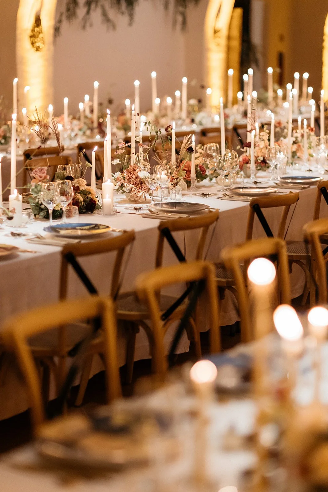 A long banquet table decorated for a wedding or special event, featuring candles, floral arrangements, glassware, and dinner plates with chairs arranged along both sides.