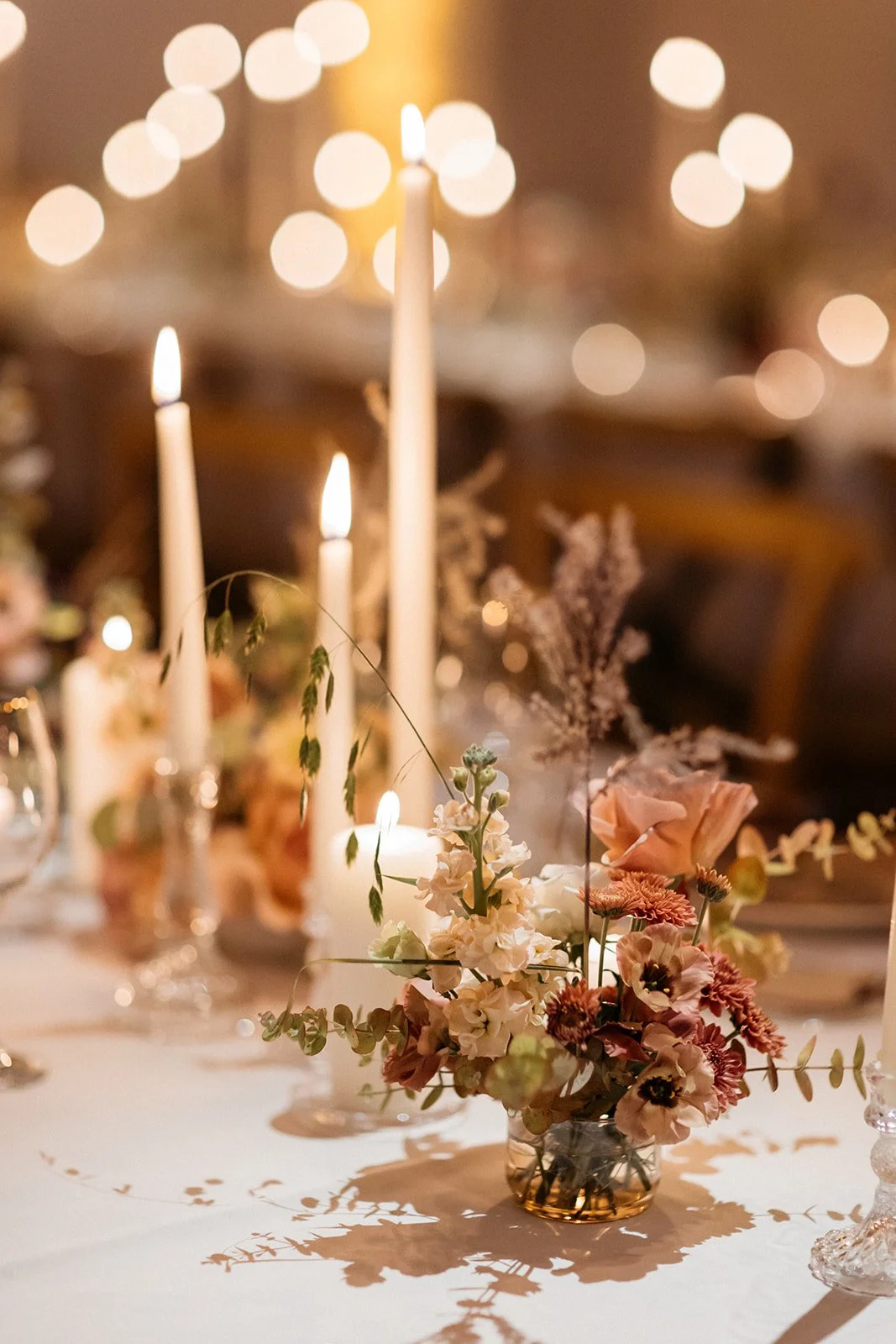 A floral centerpiece with pink and white flowers on a table, surrounded by lit taper candles and glassware, with blurred string lights in the background.
