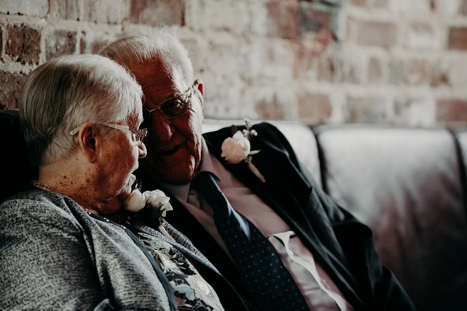 An elderly man and woman sitting closely together on a bench against a brick wall, wearing formal attire with corsages, sharing a tender moment.