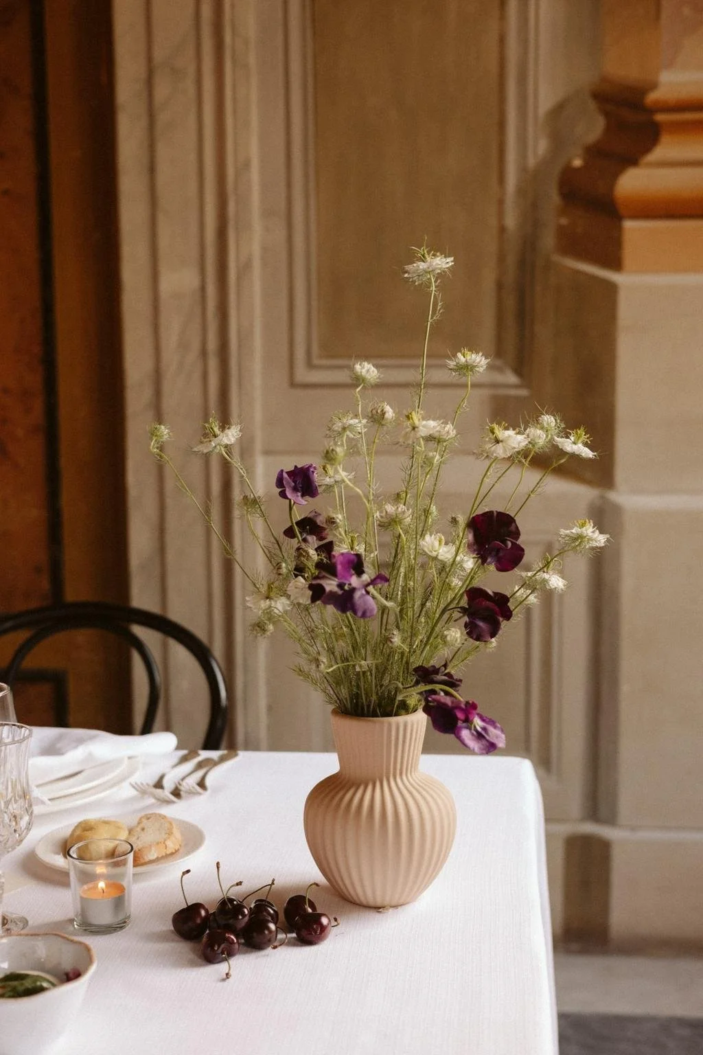 A beige vase with ribbed design holding a bouquet of white and purple flowers on a white tablecloth, with cherries, a lit candle, and a glass on a table in a room with wooden and cream-colored paneling.