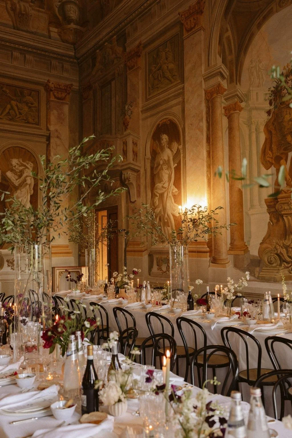 Elegant banquet table in a grand hall with classical statues, ornate wall murals, and tall vases of greenery and flowers, set for a formal dinner.
