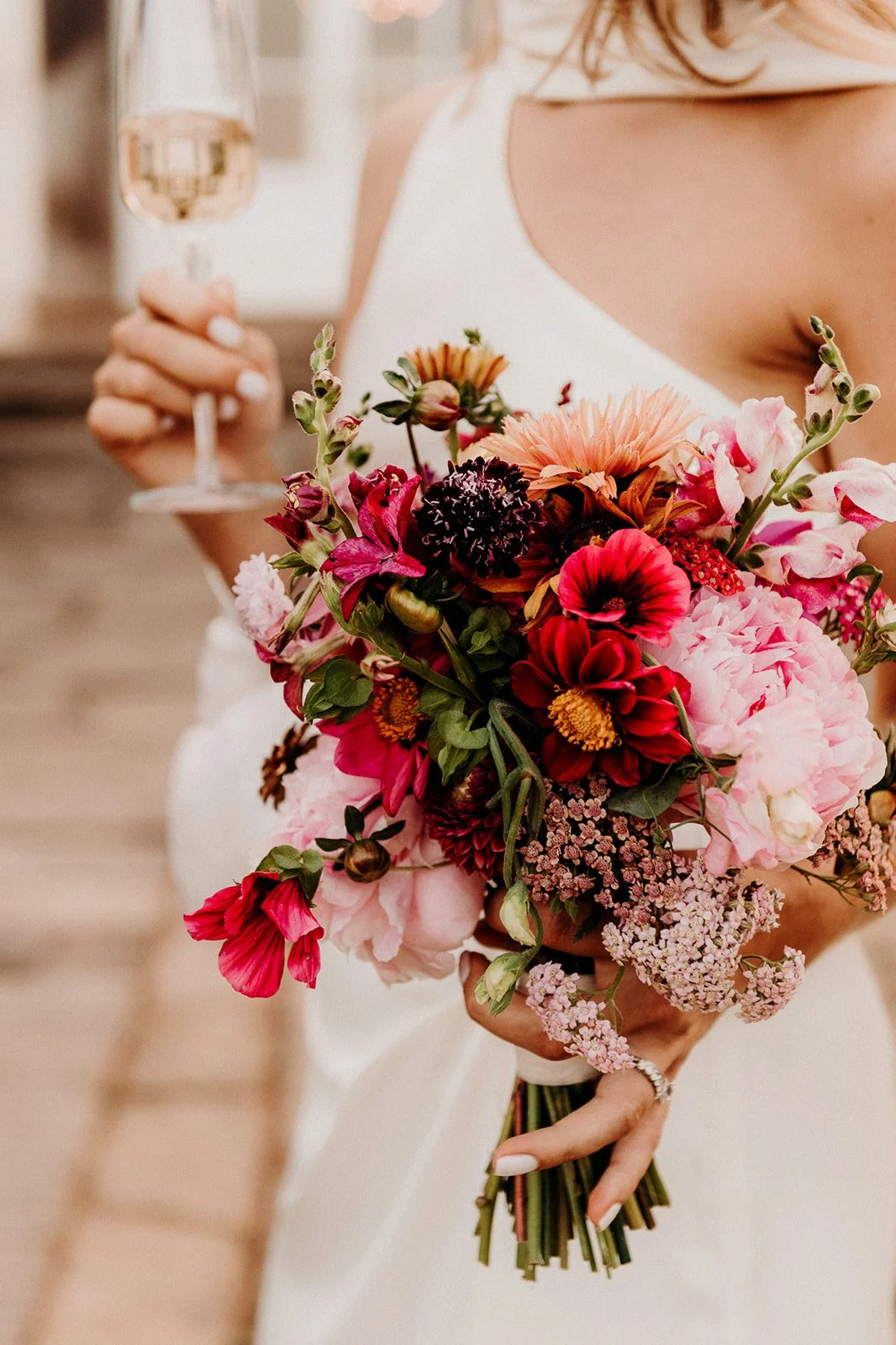 A bride holding a colorful bouquet of pink, red, and purple flowers while holding a glass of champagne.