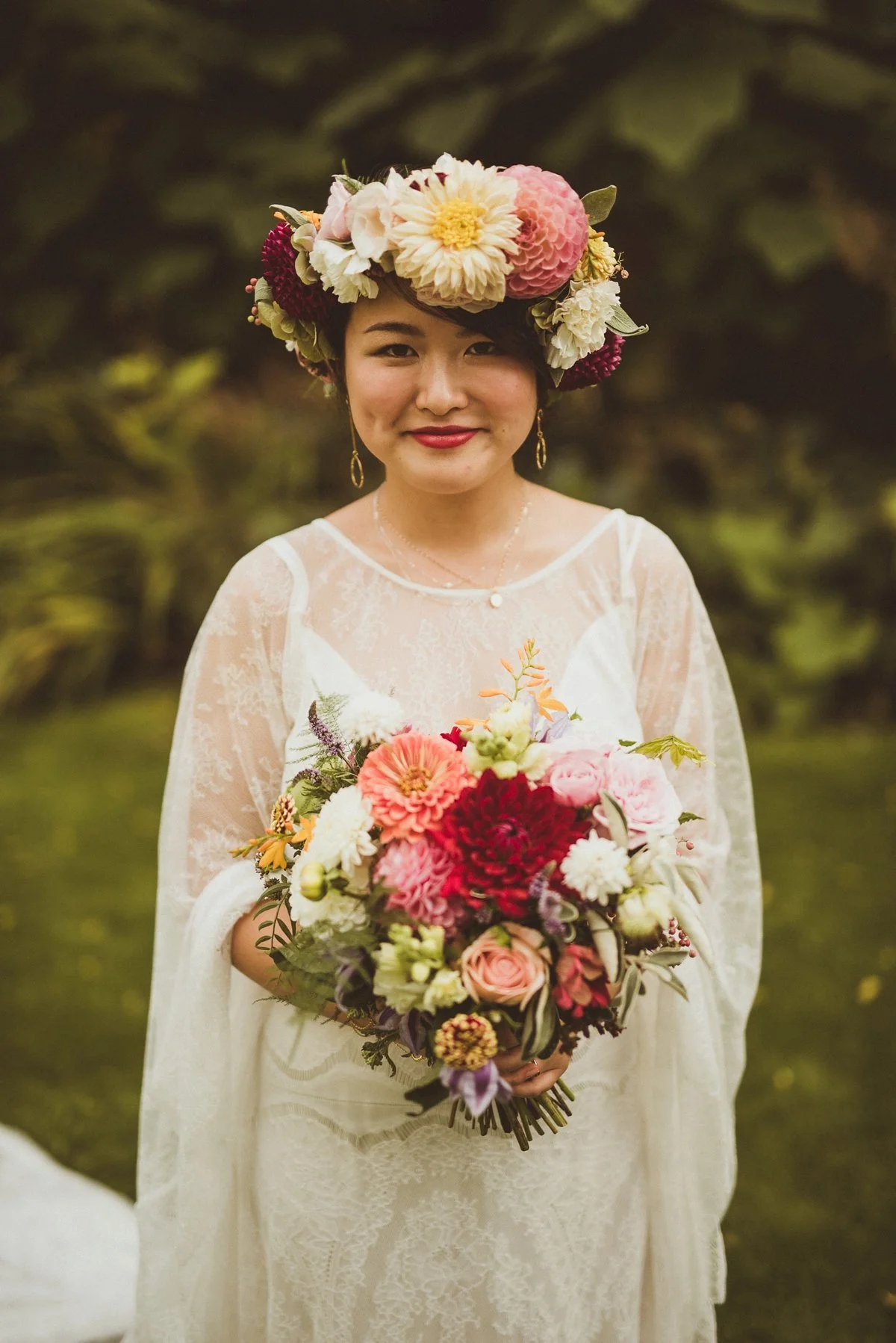 A woman with a floral crown and earrings holding a large bouquet of flowers outdoors.