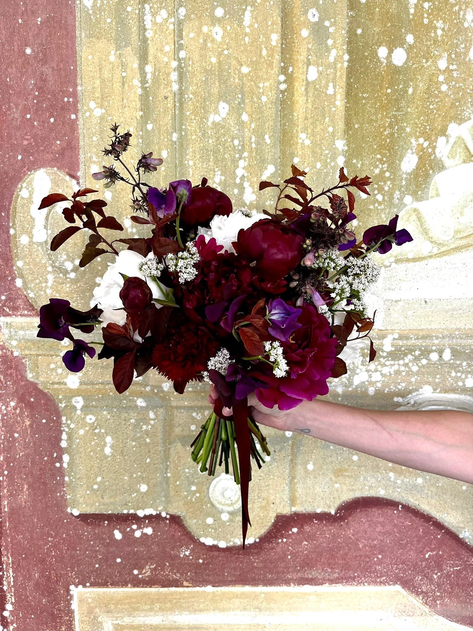 A hand holding a bouquet of mixed dark red, purple, and white flowers against a backdrop of a wall with paint splatters and graffiti.