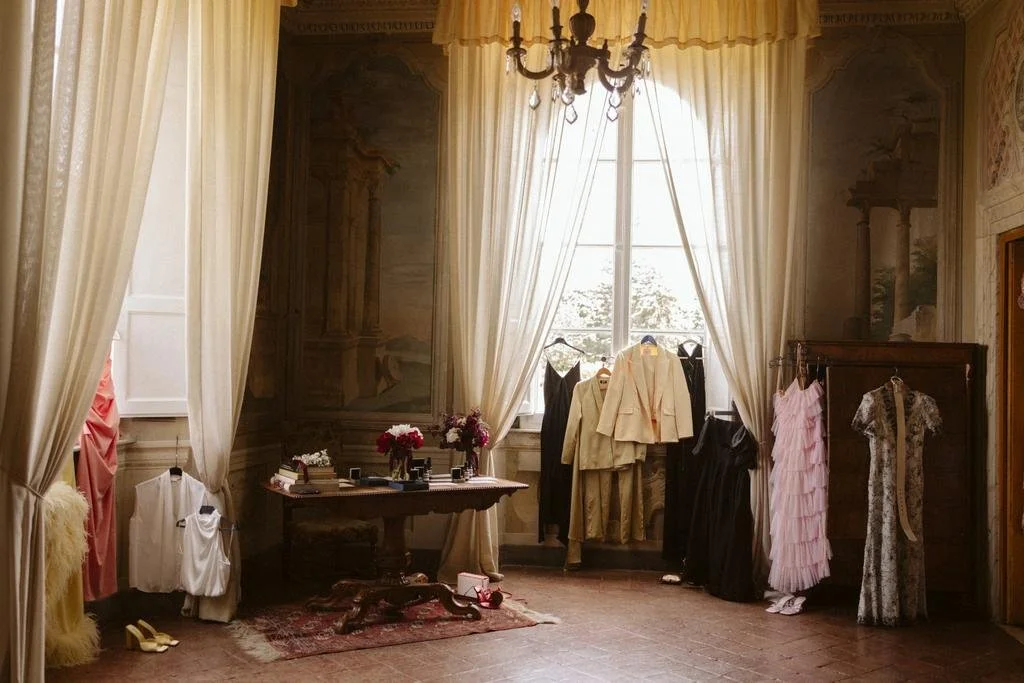 Elegant room with vintage decor, cream curtains, chandelier, wooden wardrobe, and various formal dresses and suits hanging, along with a small table with flowers and books.