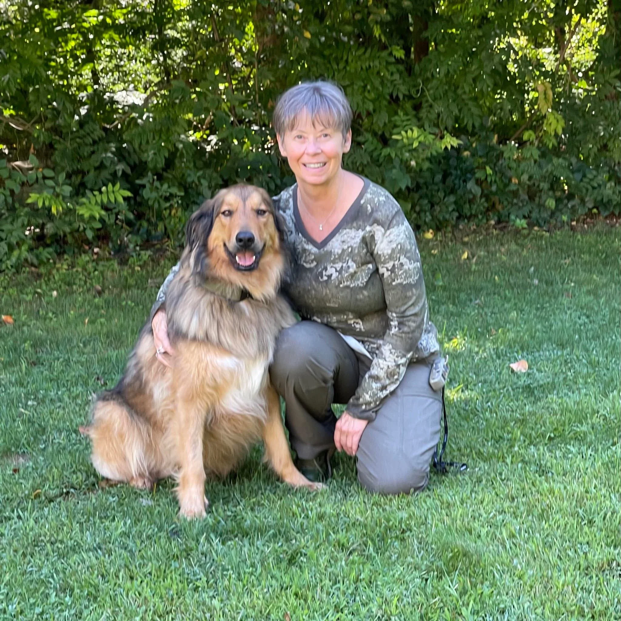 A woman with short gray hair kneeling on grass, smiling, next to a fluffy, medium-sized dog. They are outdoors with green bushes and leaves in the background.
