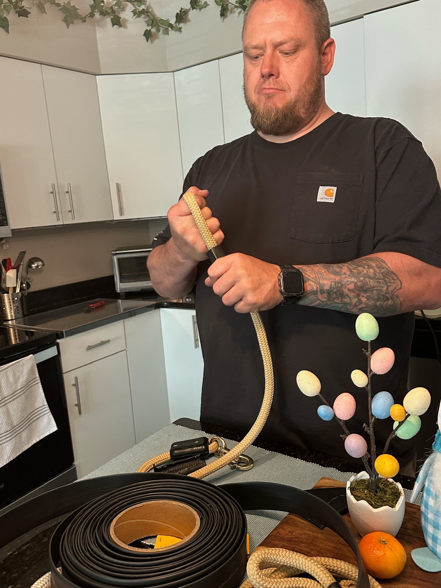 Man with tattoos on his right arm holding a rope in a kitchen. There is a small colorful Easter tree in a white pot and an orange on the table in front of him.