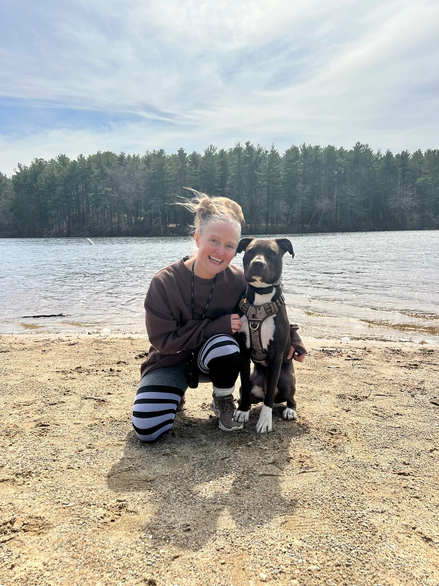 A woman smiling, kneeling on a sandy beach next to a black and white dog with a harness, with a lake and forested shoreline under a partly cloudy sky in the background.