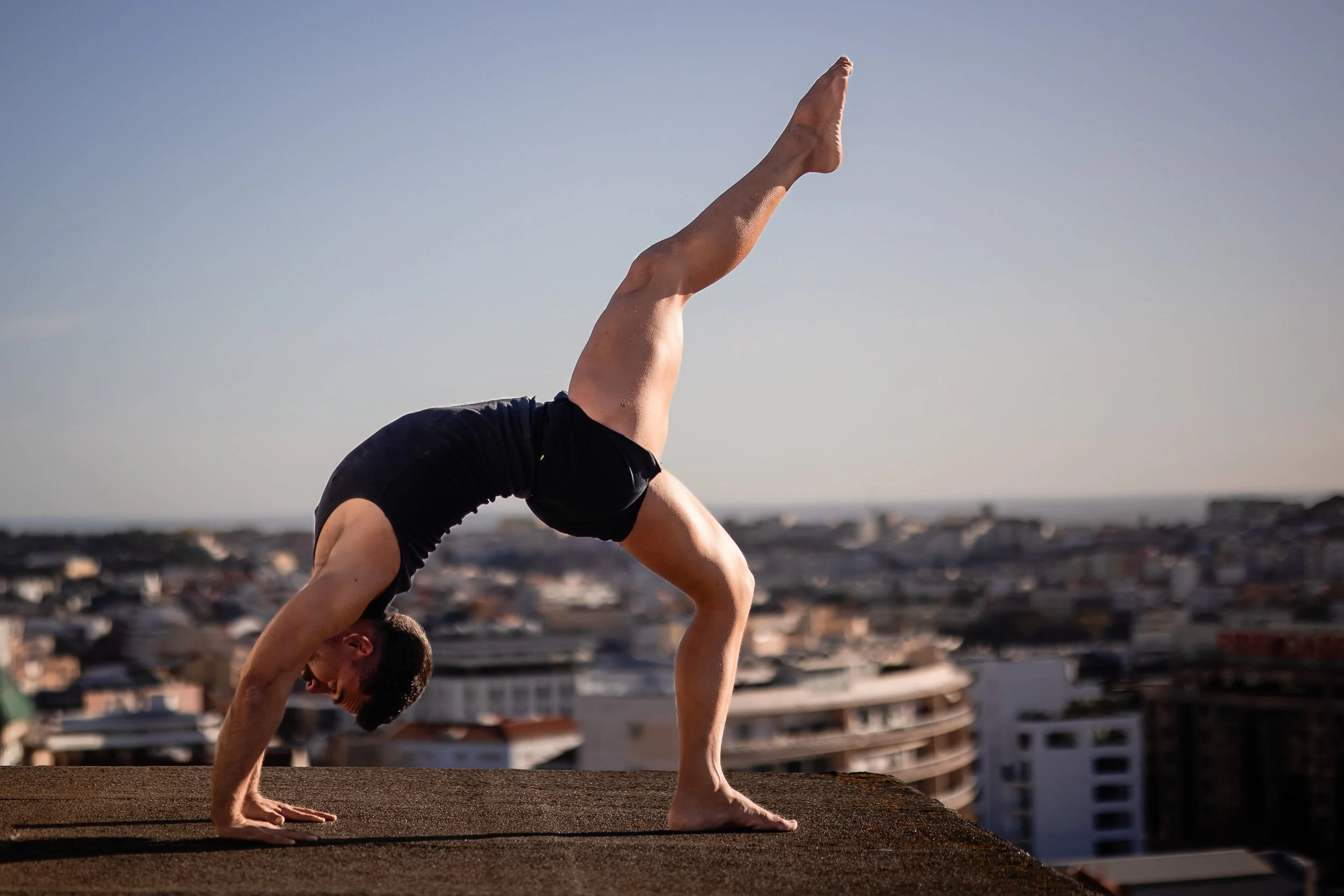 A man practicing yoga on a rooftop at sunset with a cityscape in the background, performing a forearm stand with one leg extended upward.