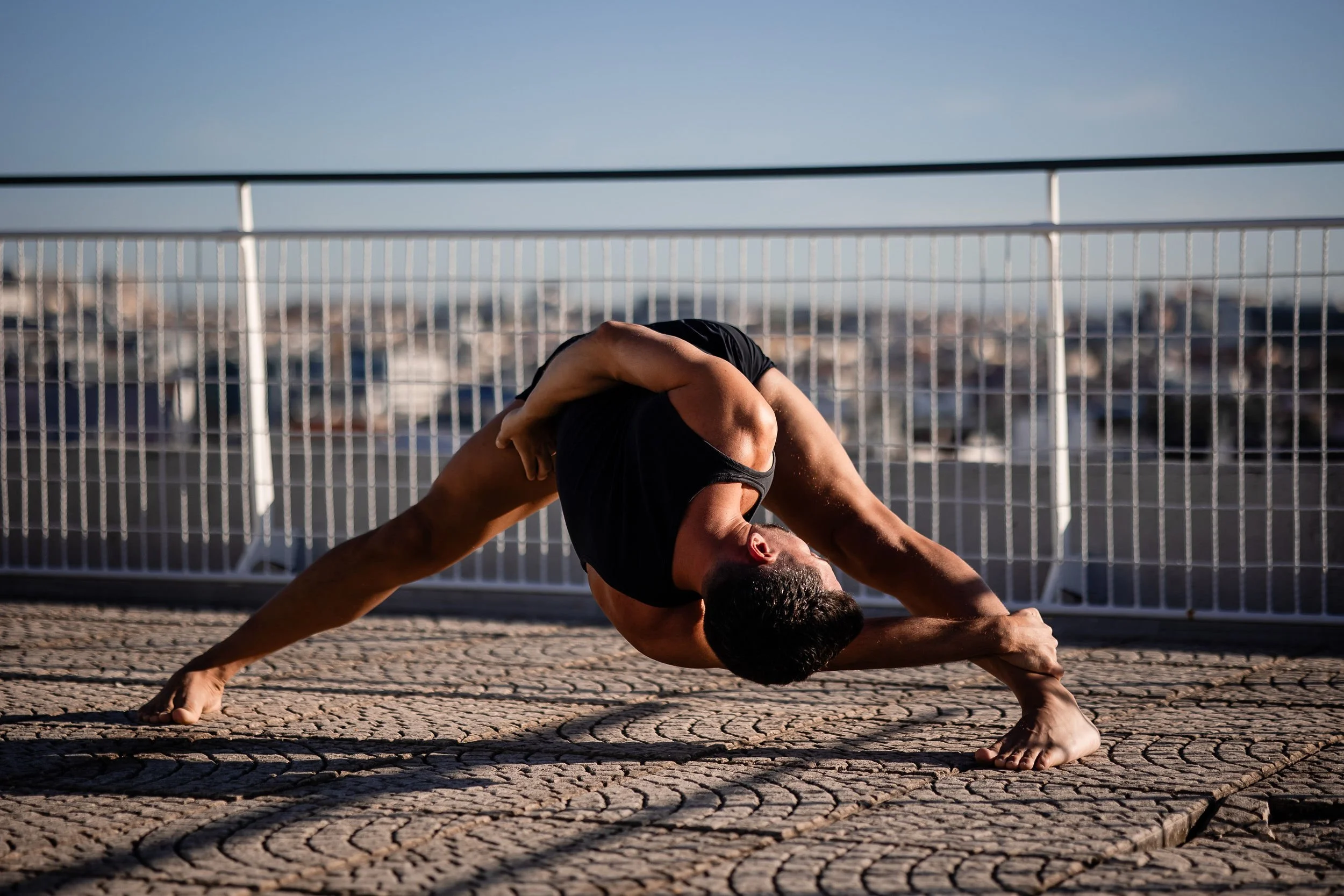 A man performing a yoga stretch outdoors on a paved surface, with a railing and cityscape in the background.