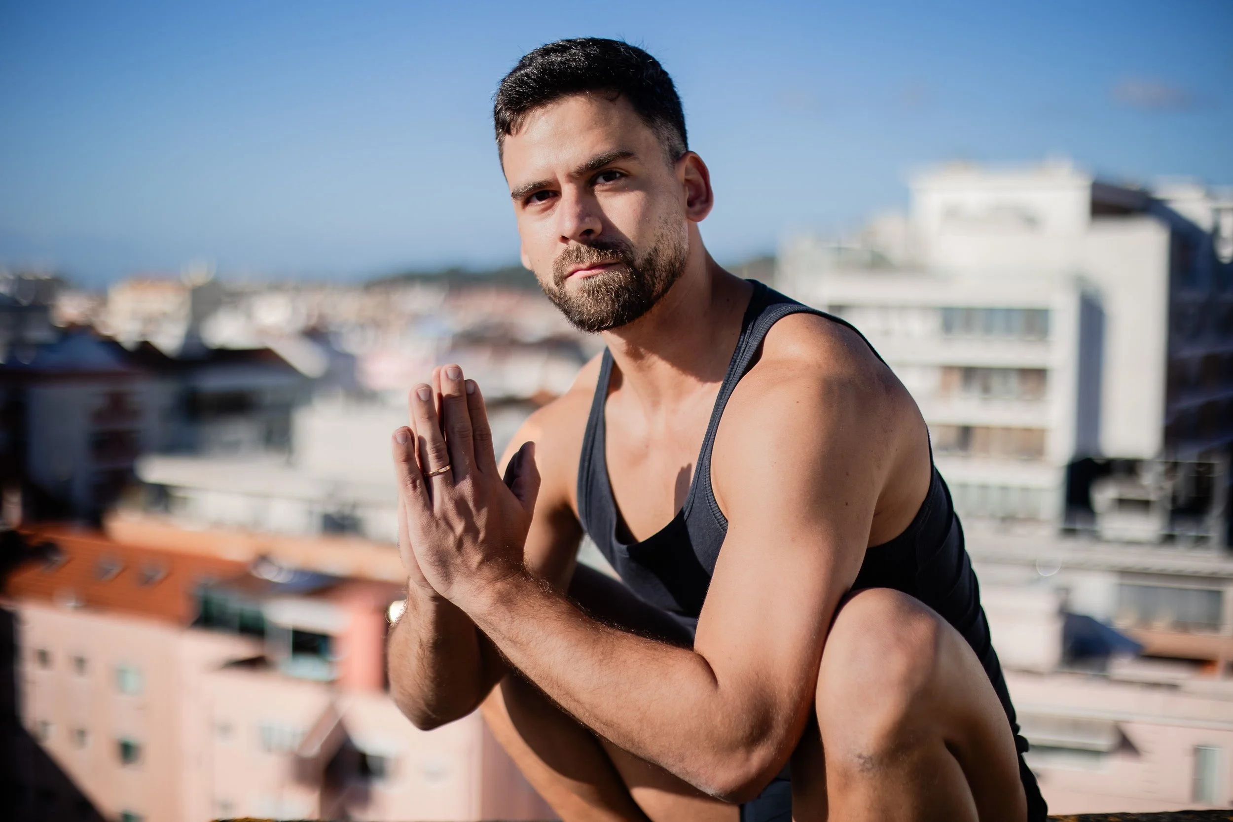 A man with a beard and short dark hair doing a yoga pose outdoors on a rooftop with a cityscape background, wearing a black tank top.
