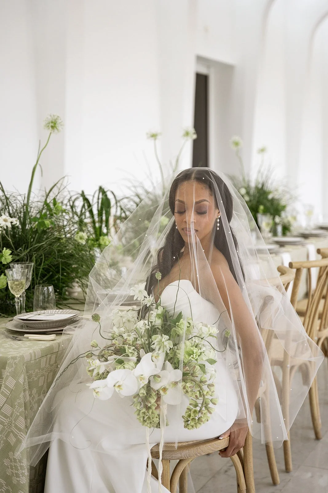 Bride sitting on a wooden chair with a veil and holding a bouquet of white flowers in a bright, modern decorated room.