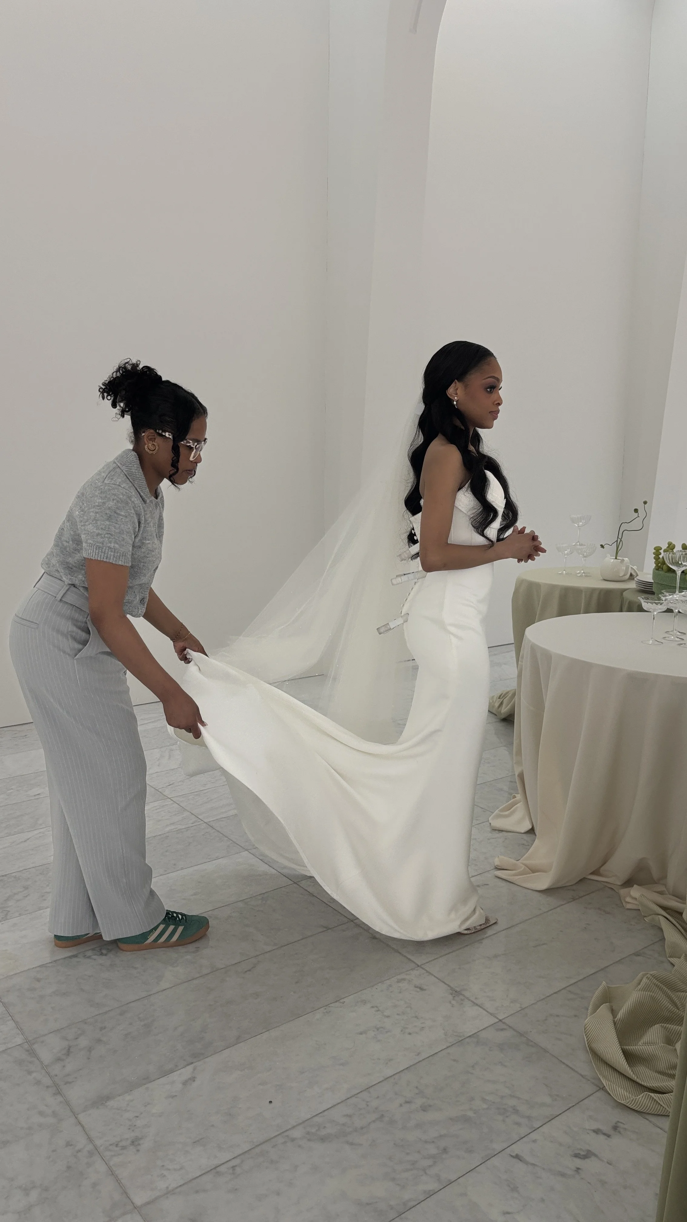 A bride in a white wedding gown having her dress adjusted by a woman in gray pants and a gray top at a wedding venue with white walls and decorated tables.