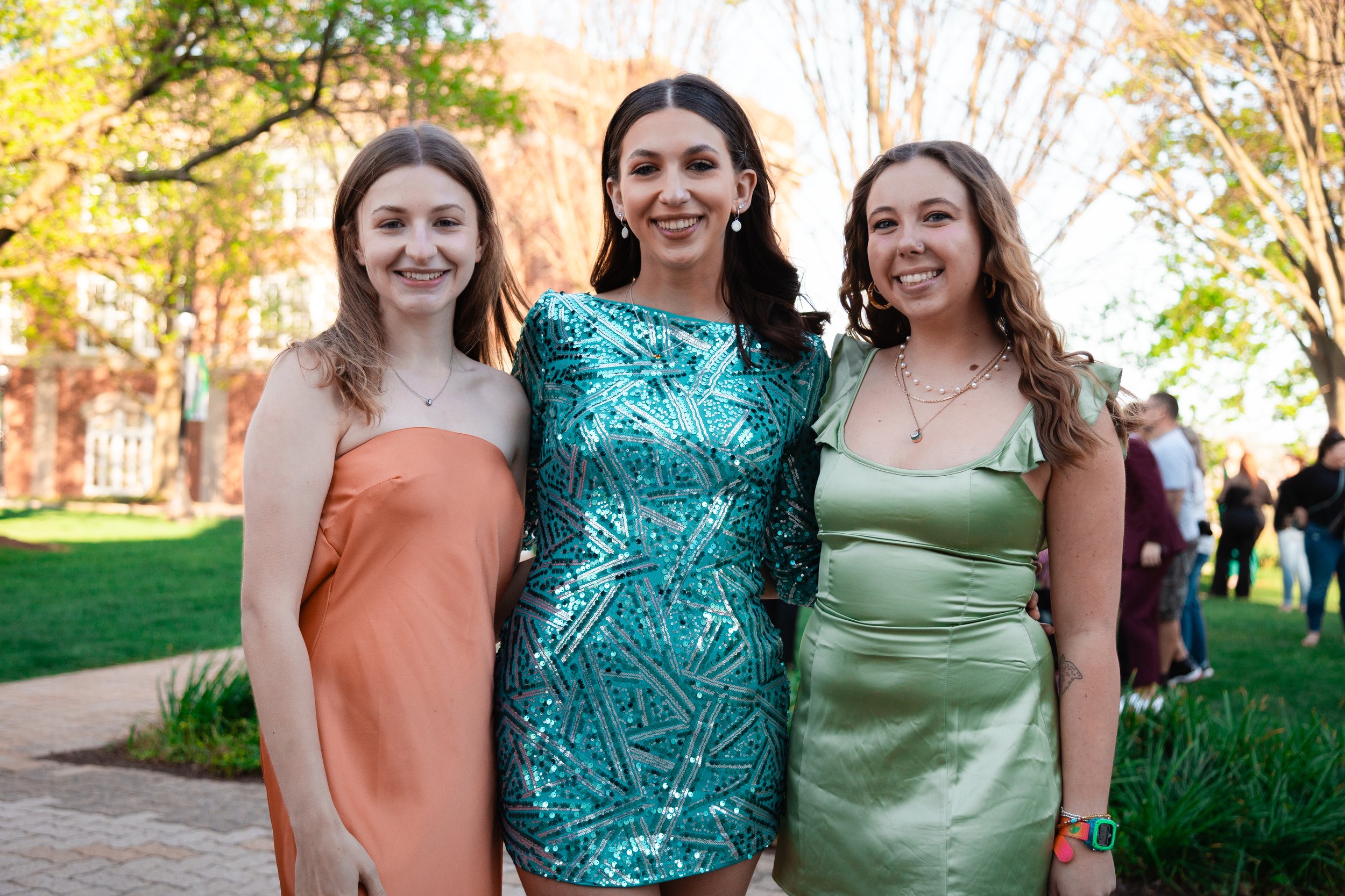 Three women posing outdoors in dresses, with trees and a building in the background.