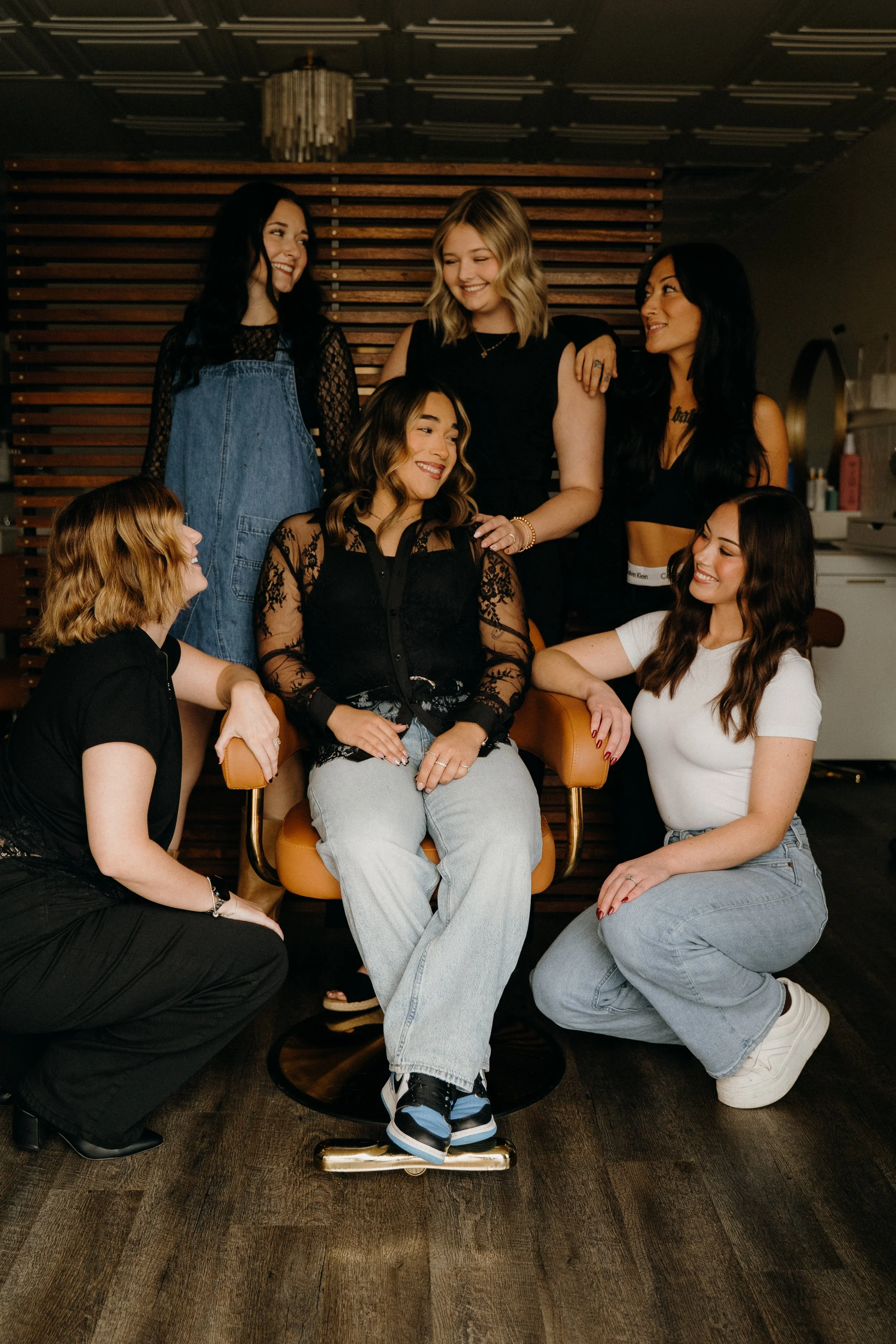 A group of seven young women with diverse hair colors and styles, smiling and interacting in a cozy indoor setting with dark wooden wall paneling.