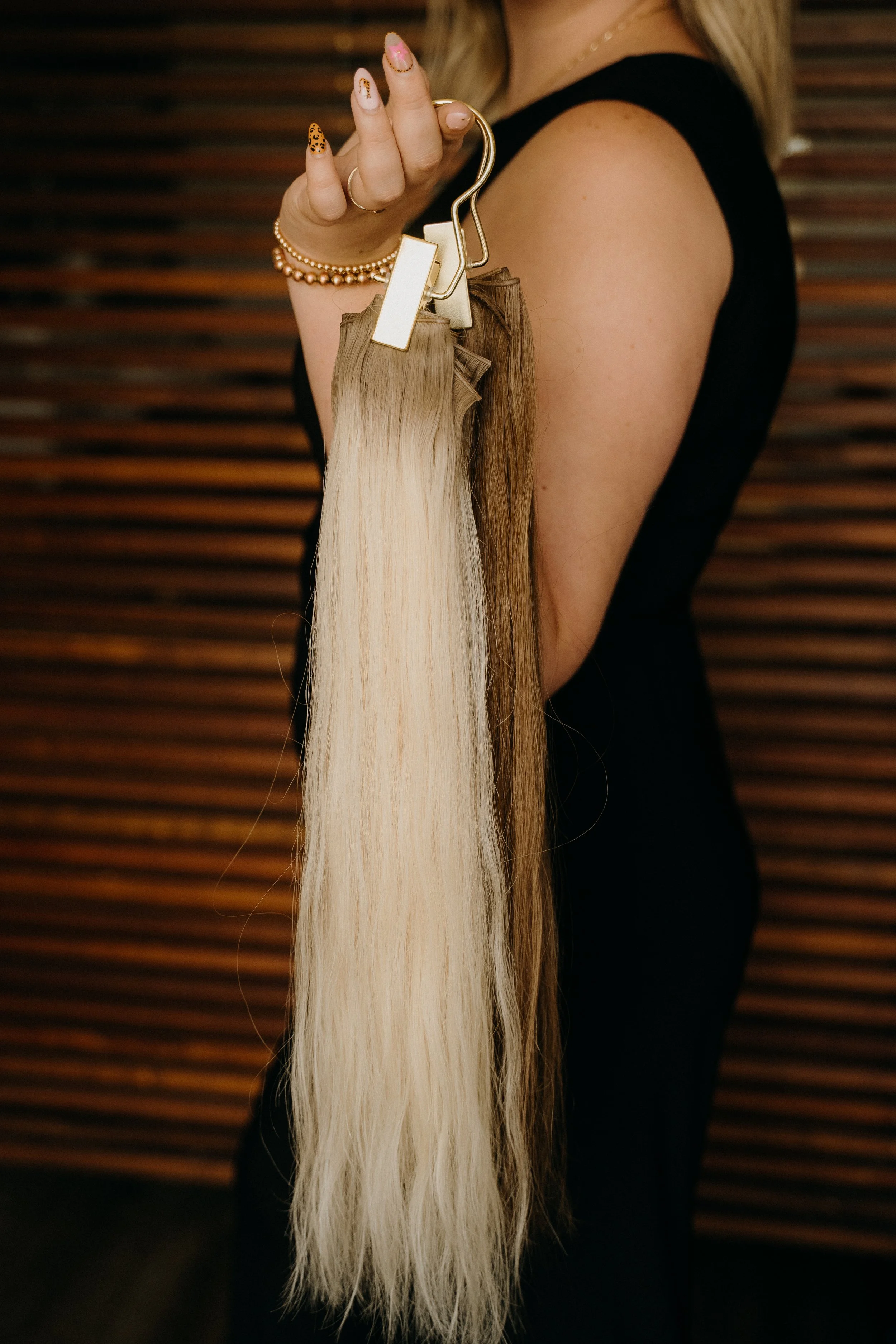 Close-up of a woman holding a hair extension with blonde hair, wearing a black sleeveless top, jewelry including a necklace, bracelet, and rings, standing in front of a wooden slatted background.