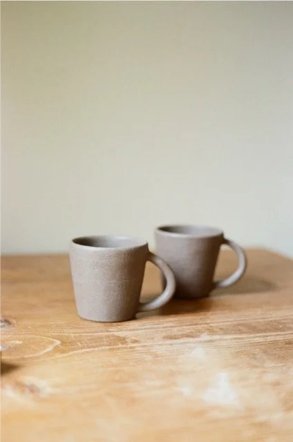 Two beige ceramic mugs on a wooden table against a plain light-colored wall.