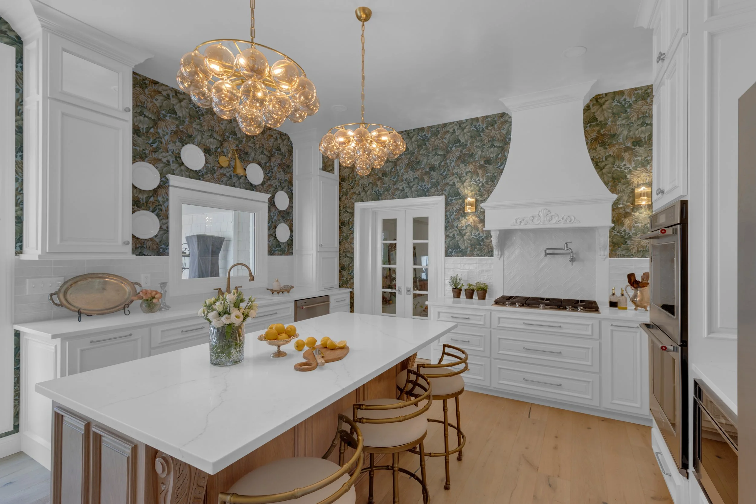 Modern kitchen with white cabinets, a large island with seating, floral wallpaper, and dual bubble chandeliers.