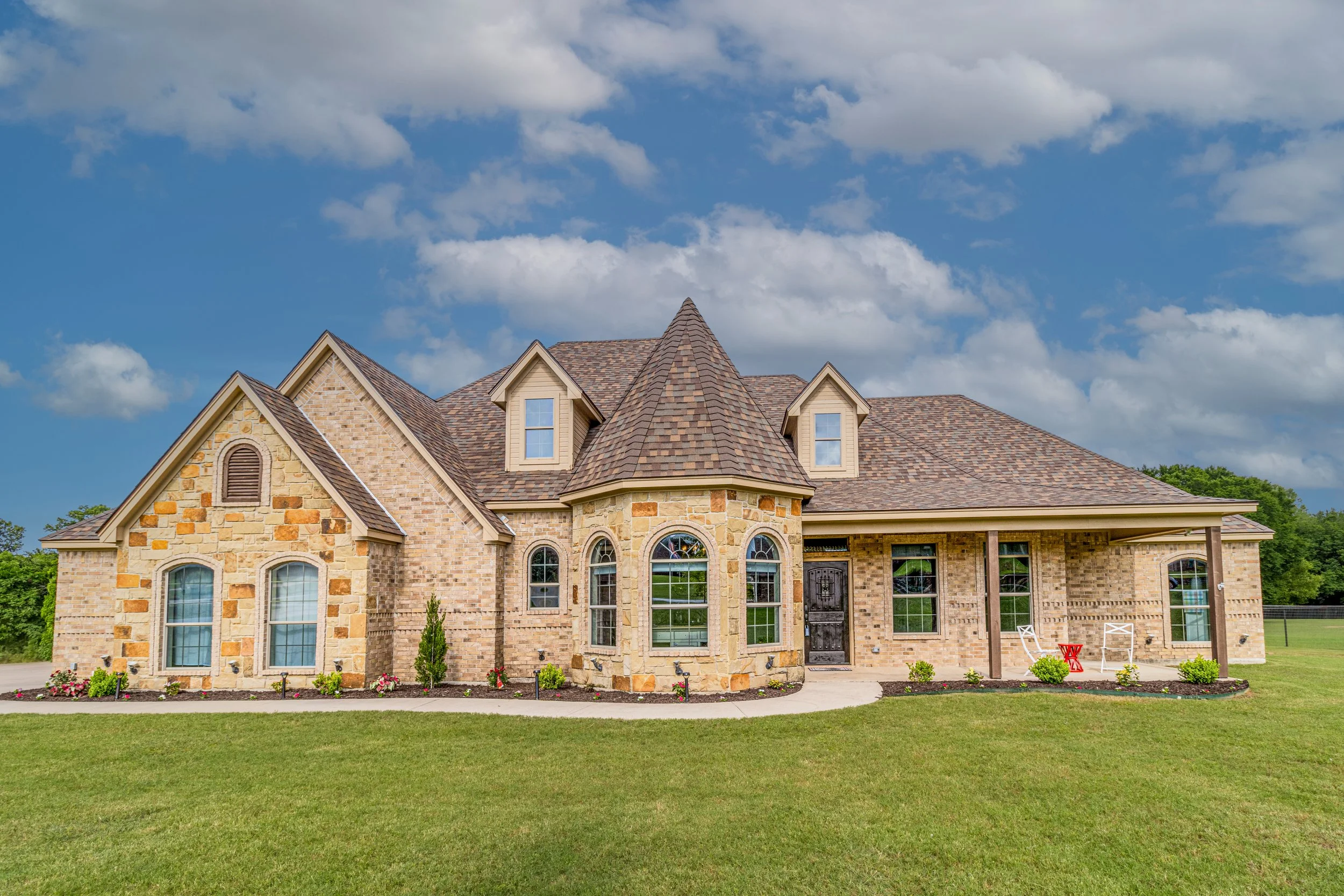 A large brick house with a landscaped front yard, multiple windows, a turret, and a porch with two chairs.